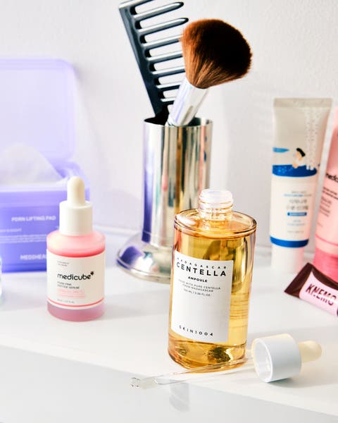 A bathroom shelf with a MEDICUBE dropper bottle, a Centella ampoule, a jar of green patches and a makeup brush.