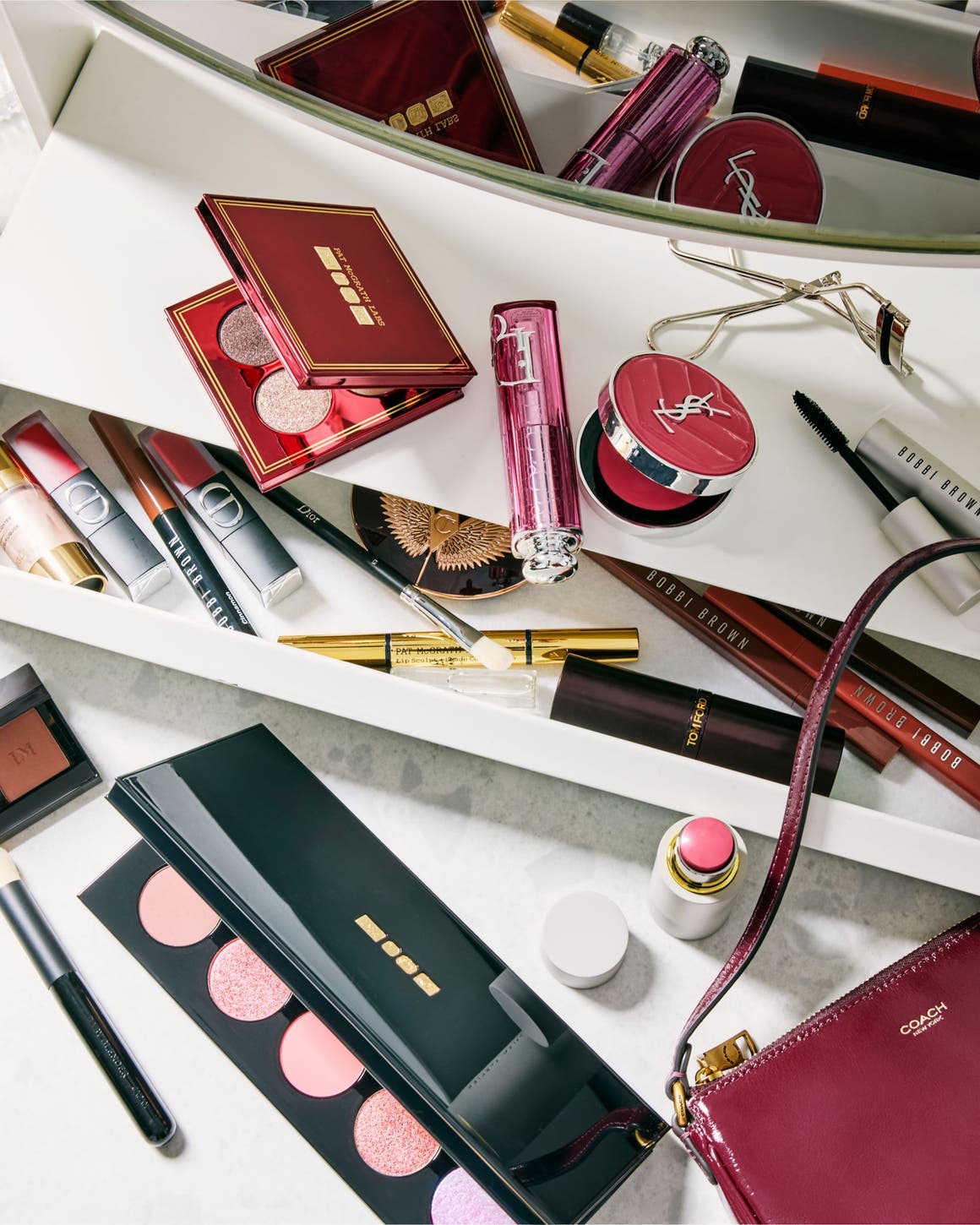 Berry-toned eyeshadow palettes, lipsticks and mascara on a white vanity beside a burgundy handbag.