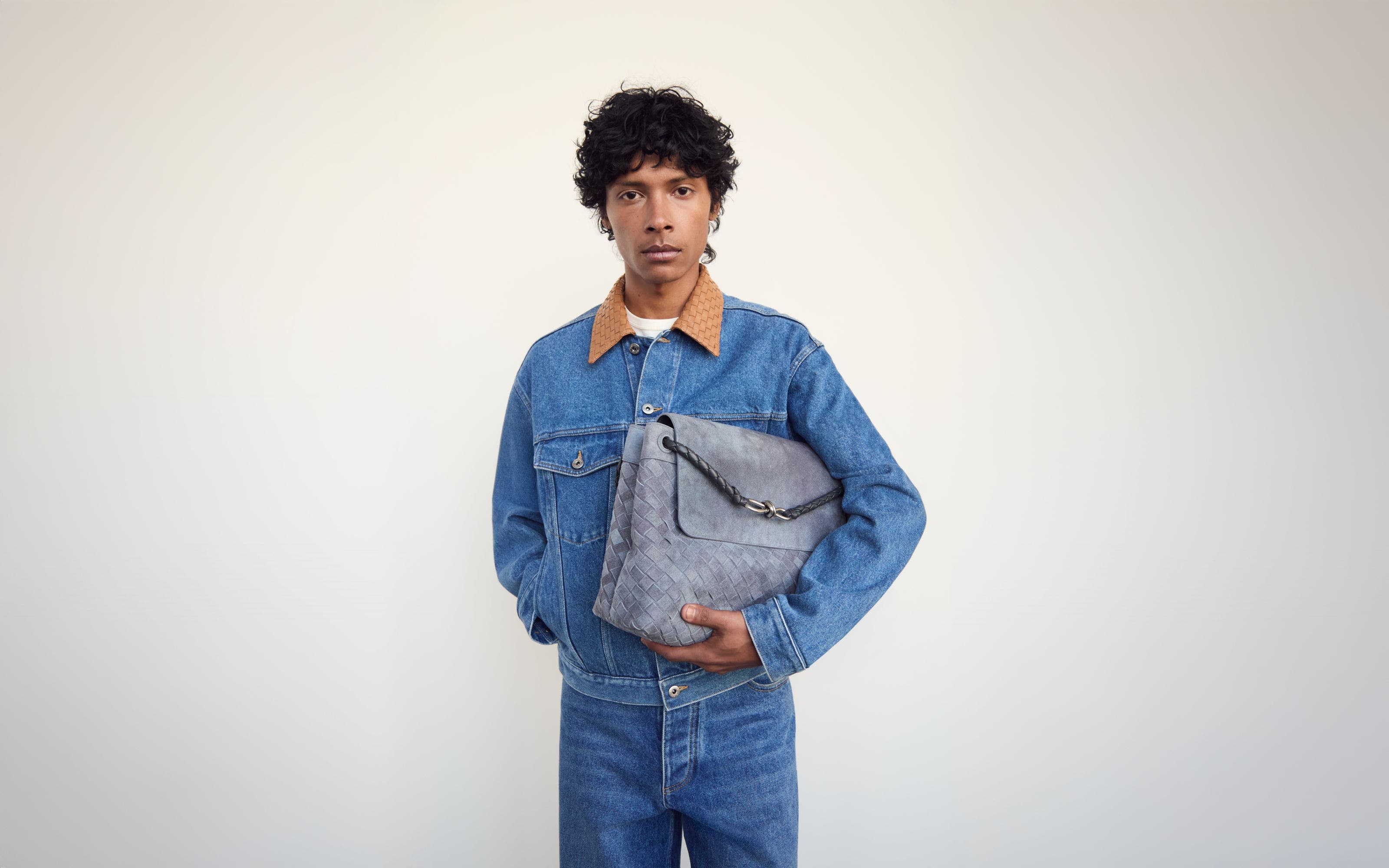 Man wearing a denim jacket and jeans and holding a woven bag.