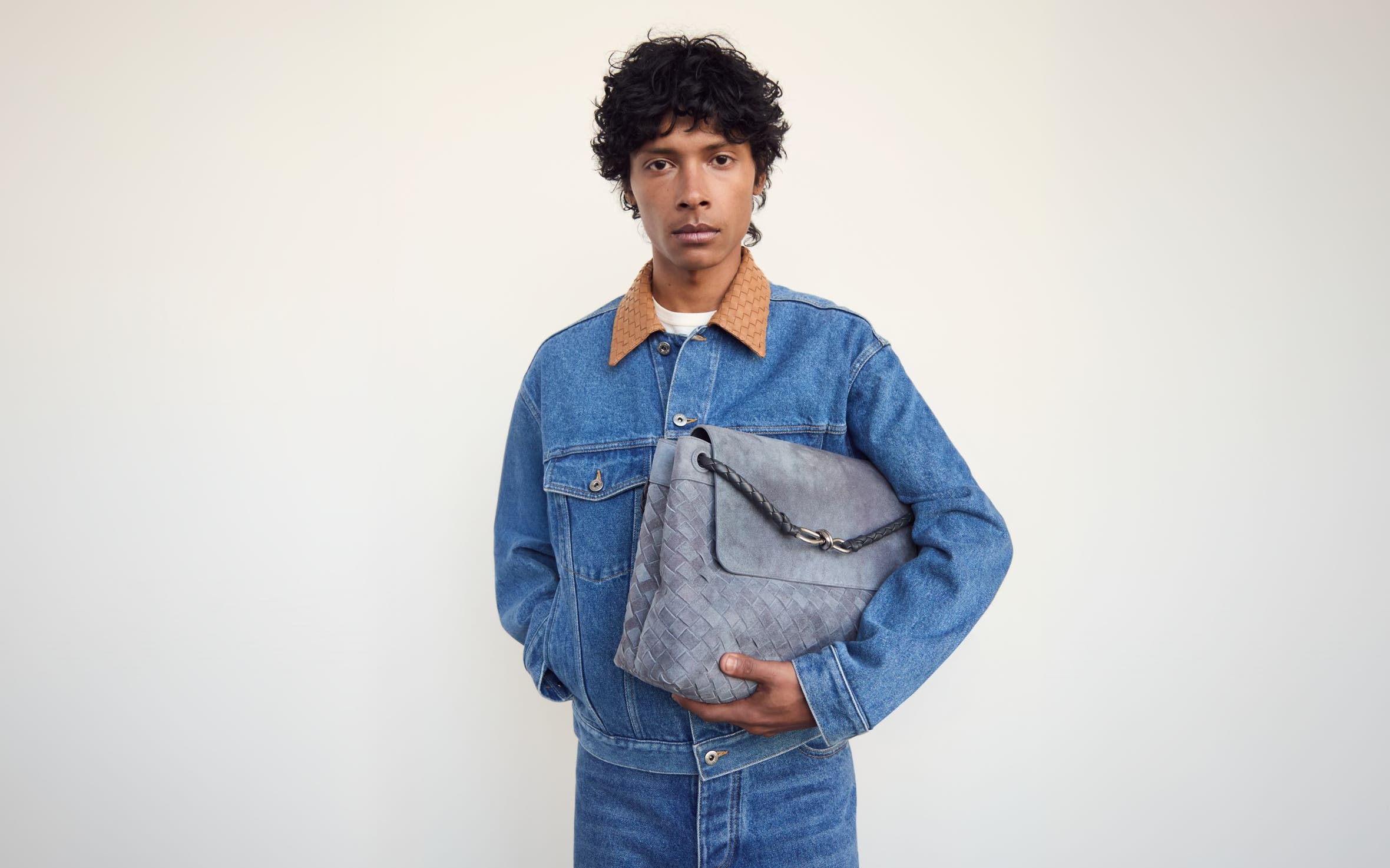 Man wearing a denim jacket and jeans and holding a woven bag.
