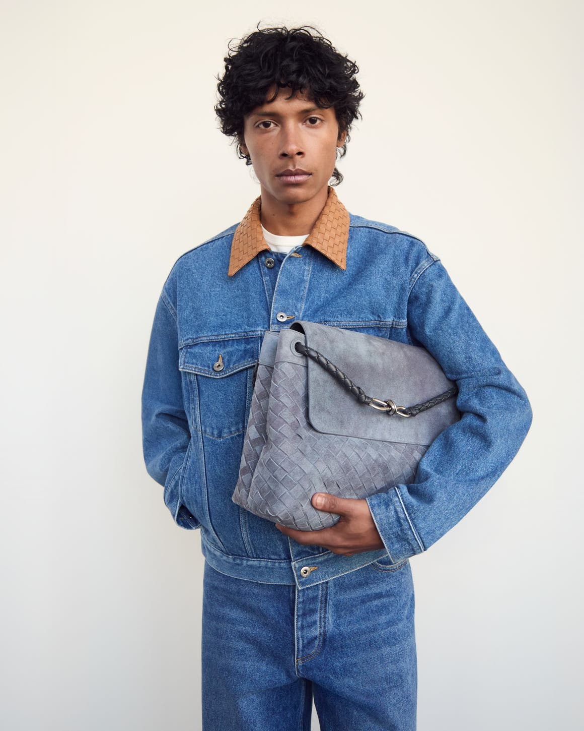 Man wearing a denim jacket and jeans and holding a woven bag.