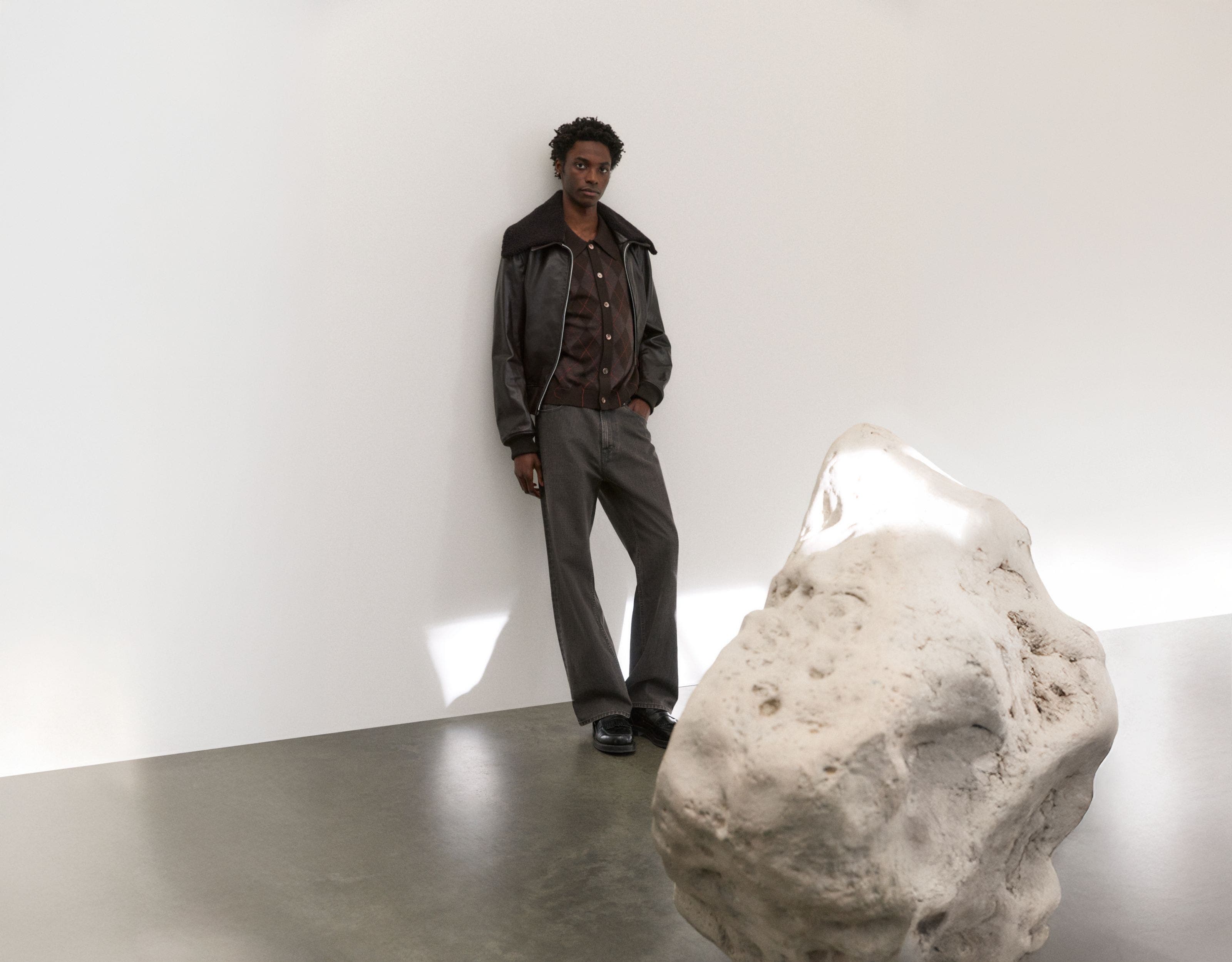 A man wearing a dark grey leather jacket, brown cardigan sweater and grey jeans.