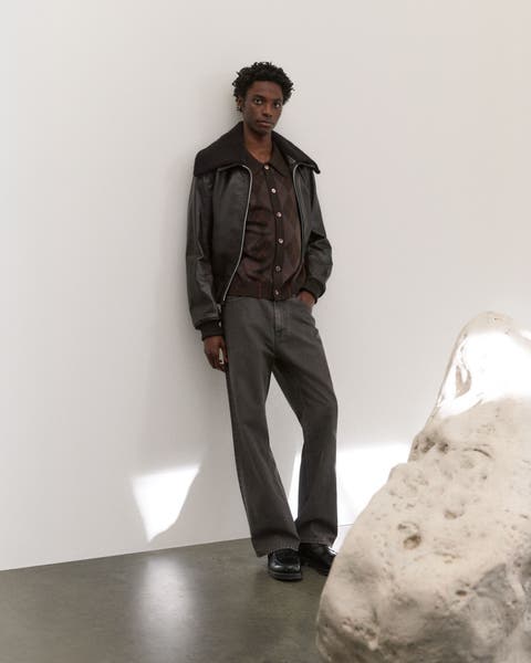 A man wearing a dark grey leather jacket, brown cardigan sweater and grey jeans.