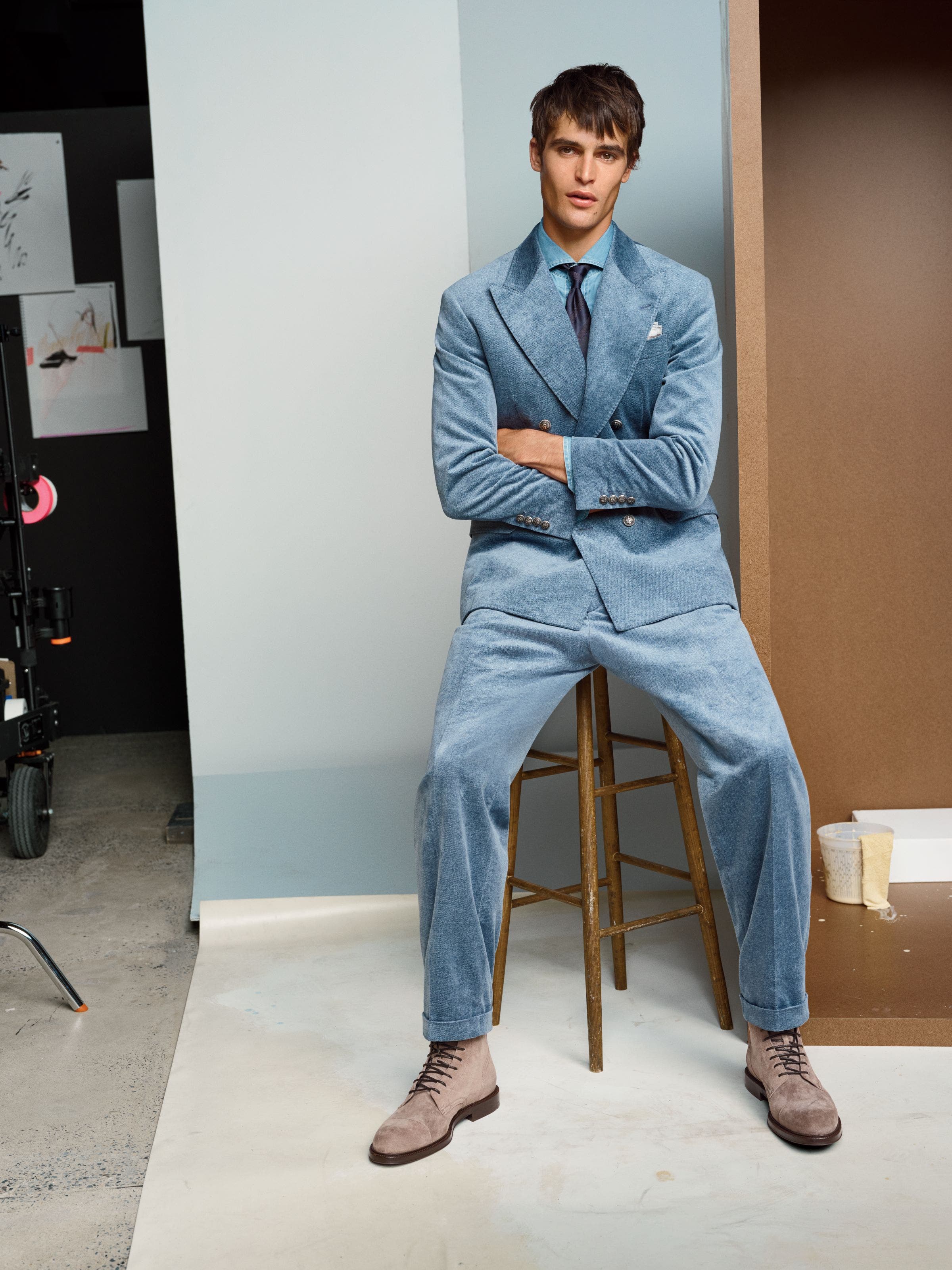Man wearing a light blue textured suit with a matching tie and pocket square, seated on a wooden stool in a photo studio