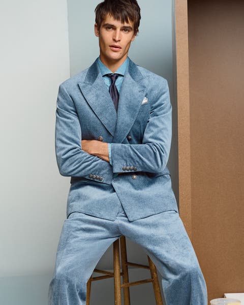 Man wearing a light blue textured suit with a matching tie and pocket square, seated on a wooden stool in a photo studio
