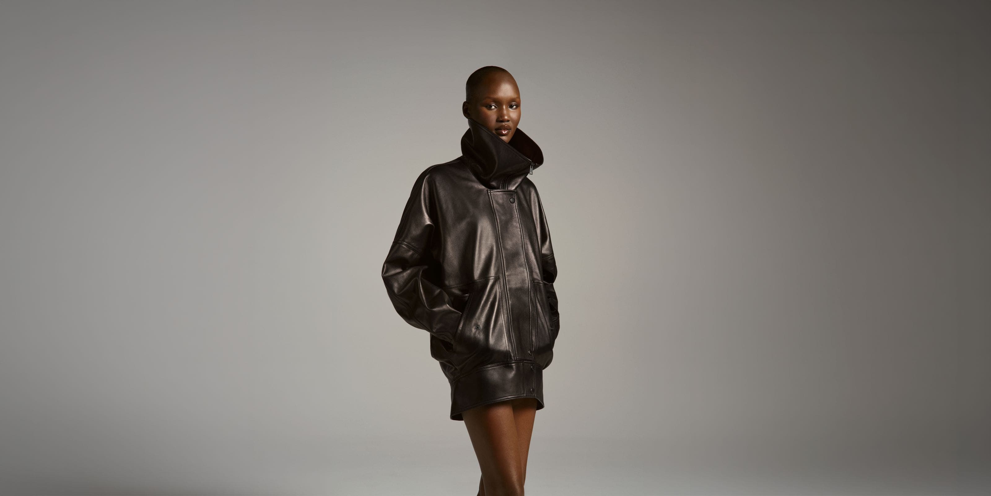 Model wearing a Balenciaga oversized dark brown leather bomber jacket with a plush shearling collar, styled with matching mini shorts and knee-high boots.