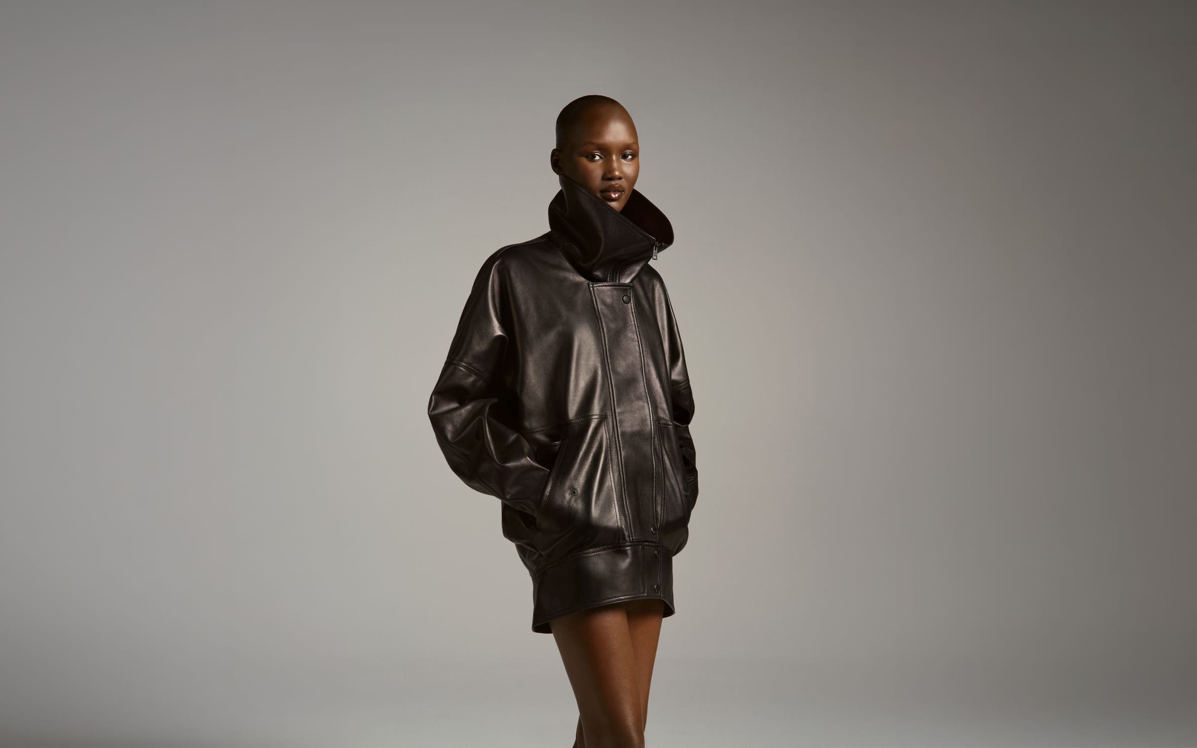 Model wearing a Balenciaga oversized dark brown leather bomber jacket with a plush shearling collar, styled with matching mini shorts and knee-high boots.