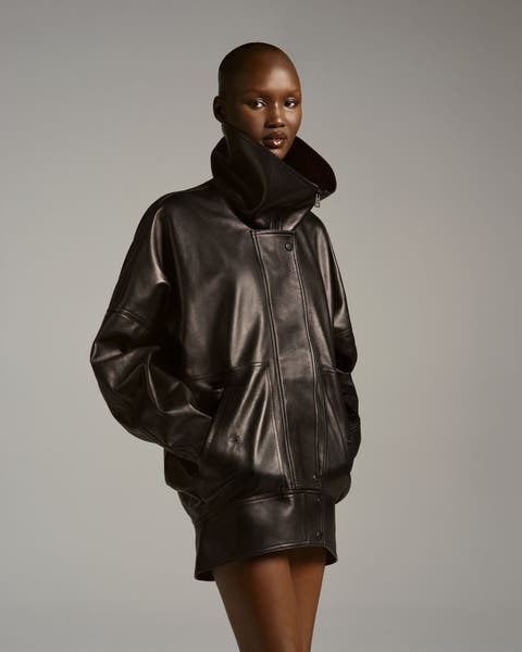 Model wearing a Balenciaga oversized dark brown leather bomber jacket with a plush shearling collar, styled with matching mini shorts and knee-high boots.