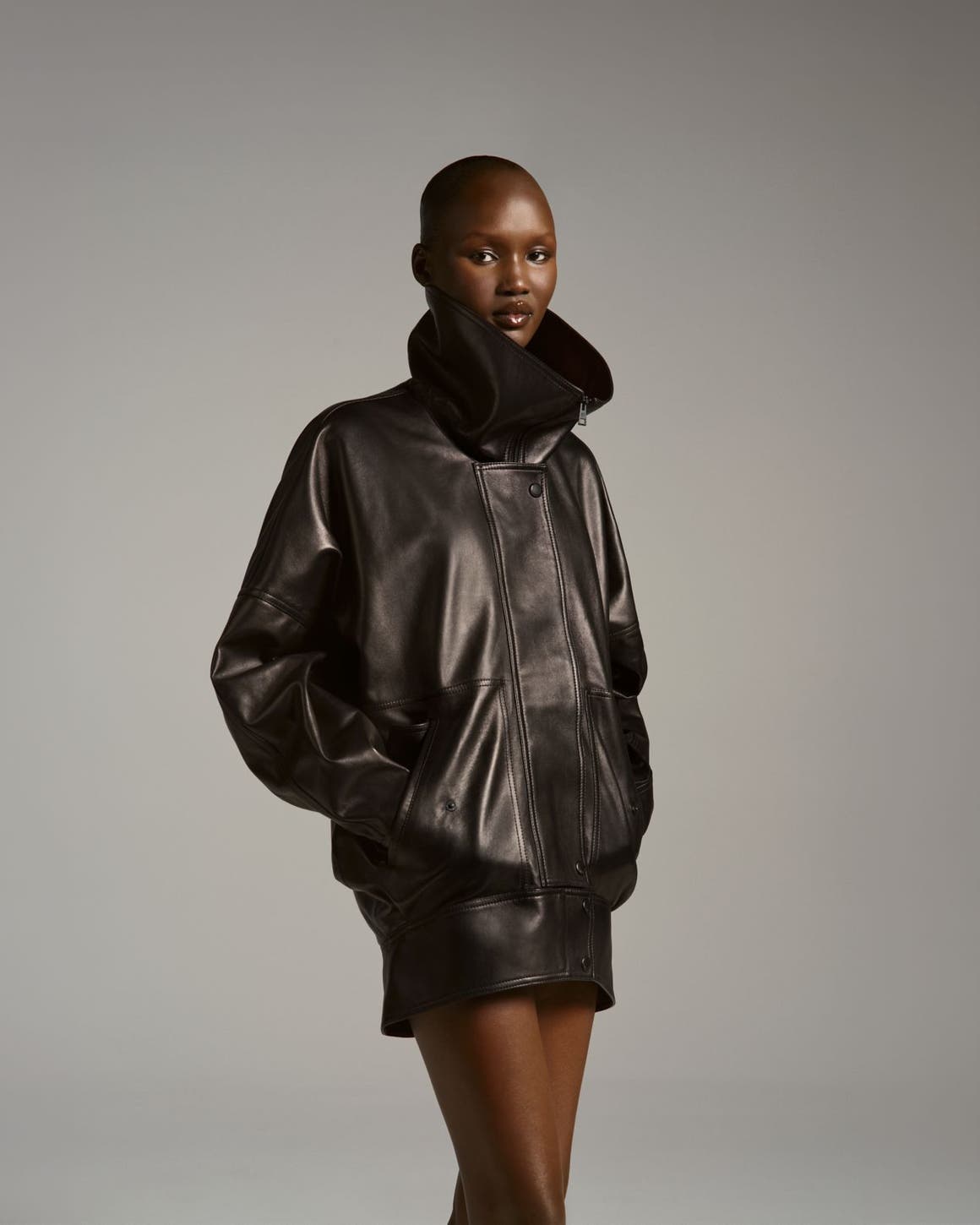 Model wearing a Balenciaga oversized dark brown leather bomber jacket with a plush shearling collar, styled with matching mini shorts and knee-high boots.