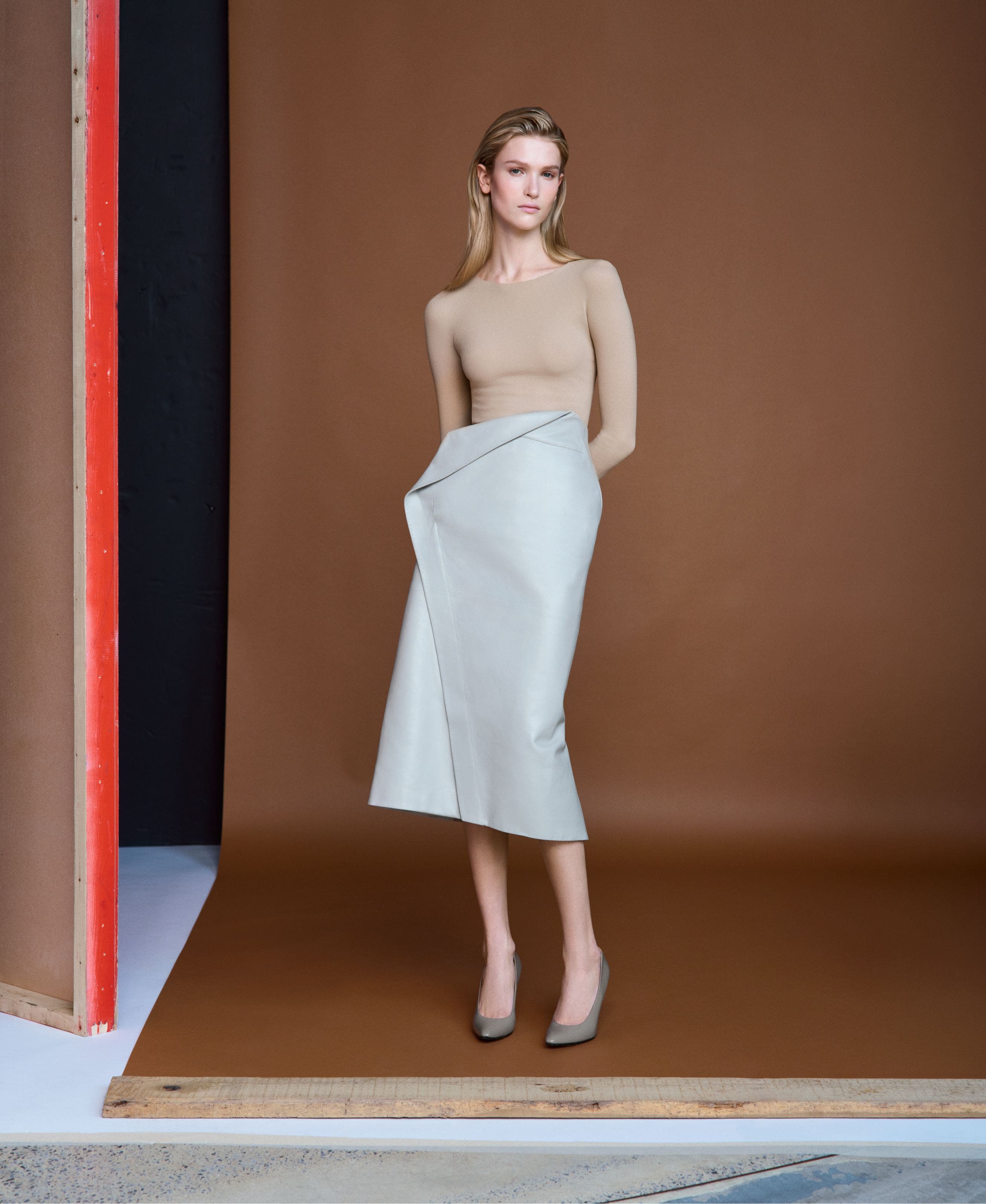 Woman wearing a beige long-sleeve top with a light blue-green wrap-style midi skirt and grey pointed-toe heels, standing against a brown backdrop