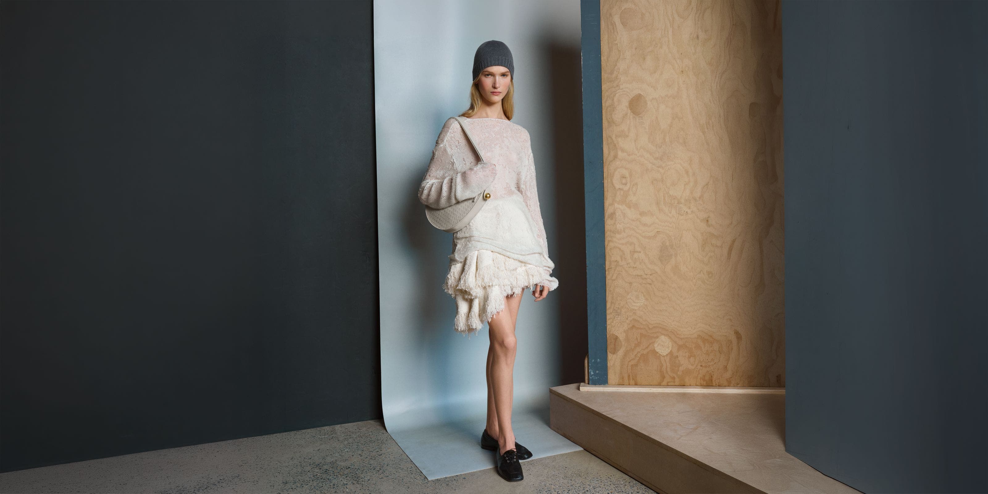 Woman in a grey beanie wearing a cream sweater and fringed white skirt, with a beige bag and black lace-up shoes.