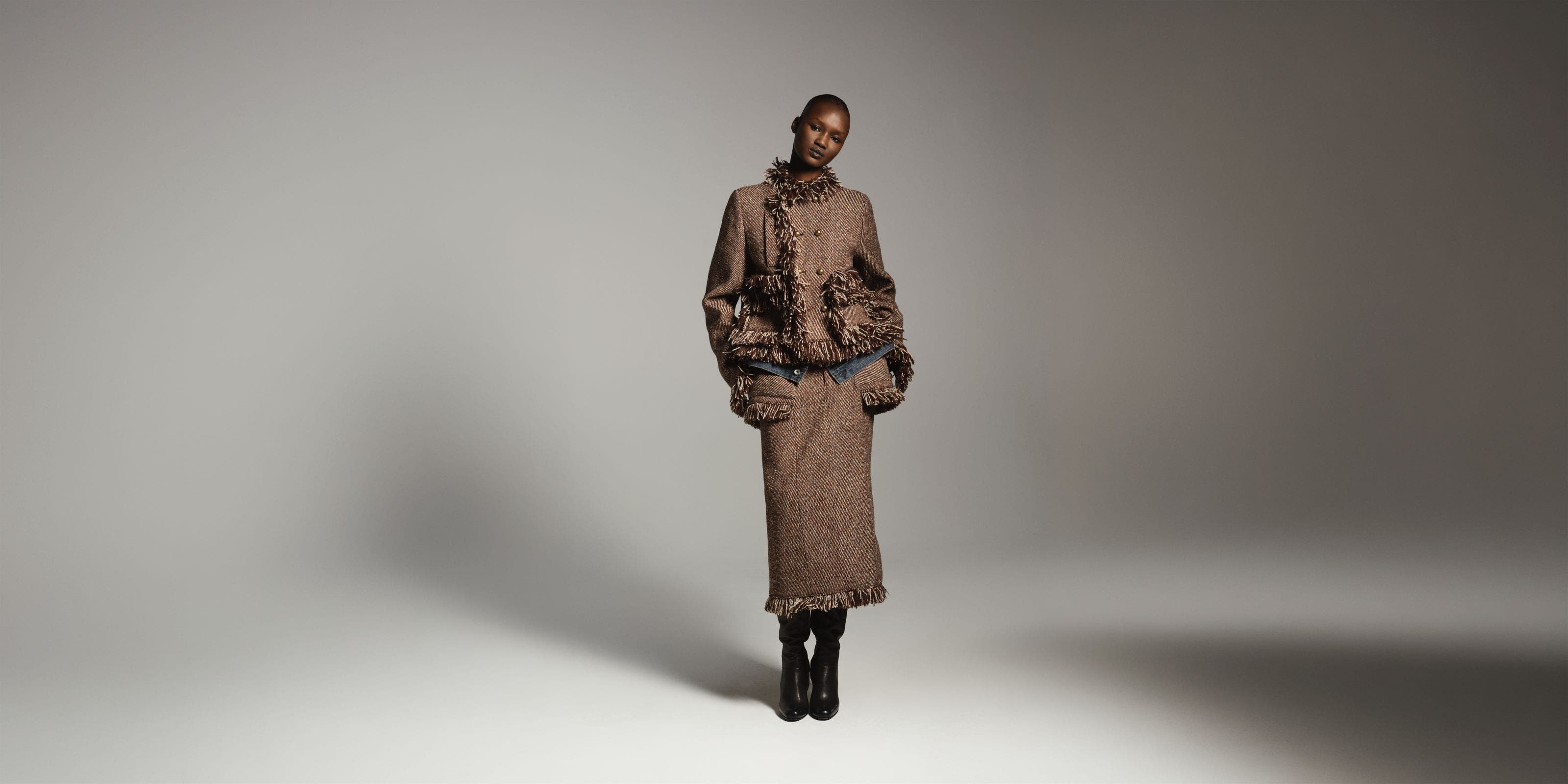 Woman wearing a textured brown fringed jacket with a matching long skirt and tall black boots, standing in a dramatically lit studio setting