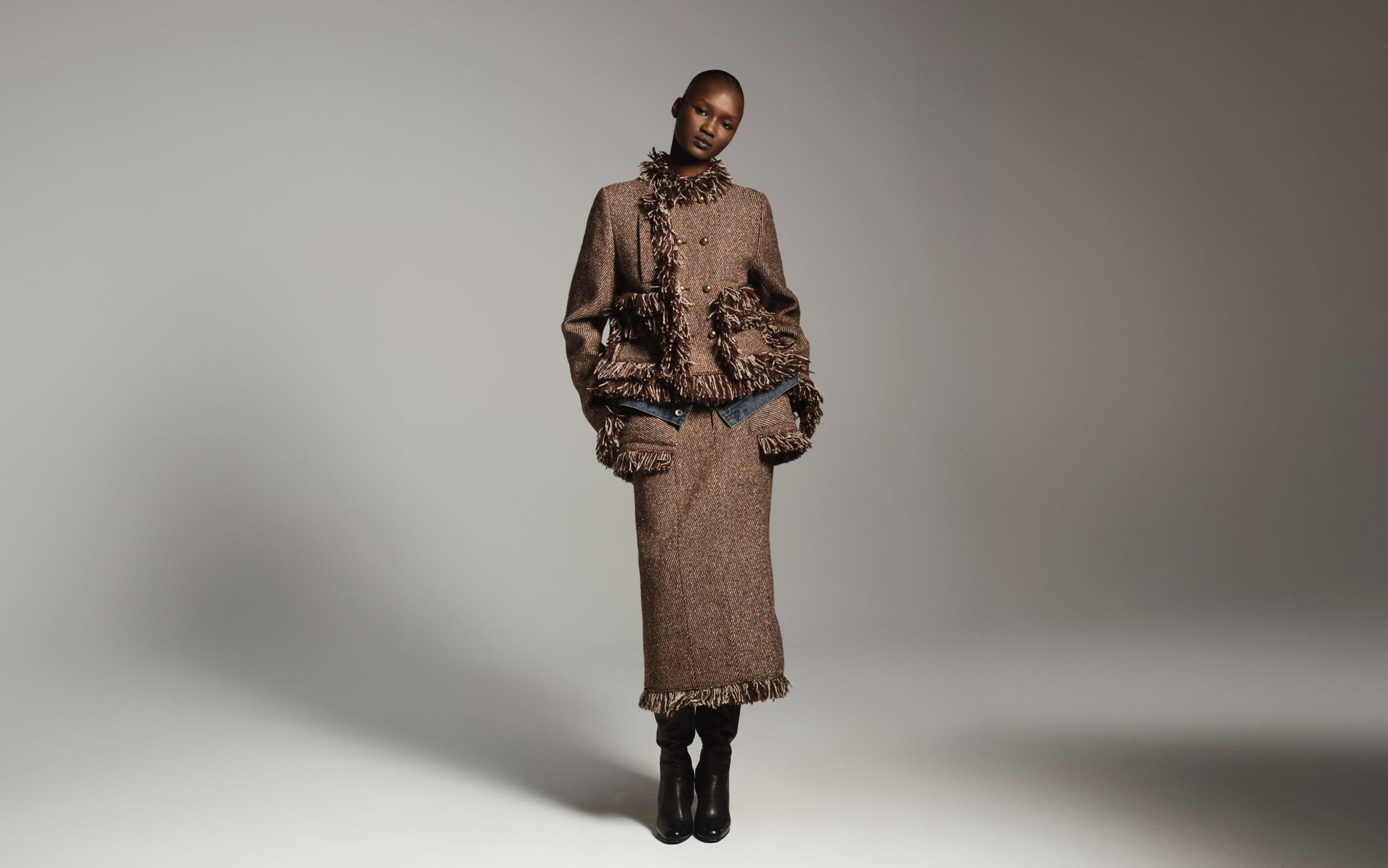 Woman wearing a textured brown fringed jacket with a matching long skirt and tall black boots, standing in a dramatically lit studio setting