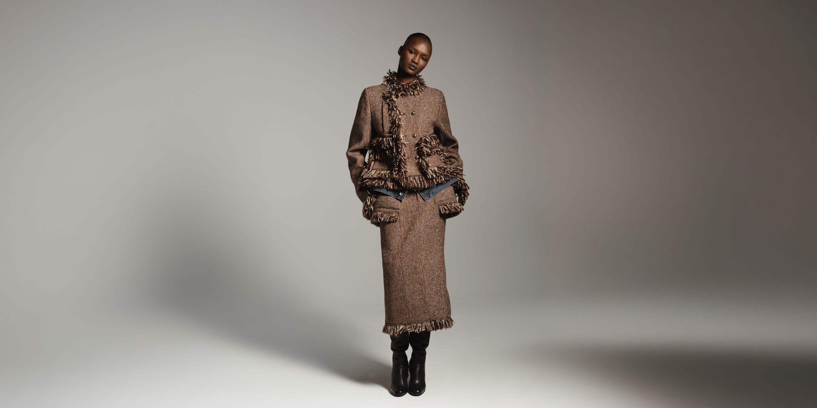 Woman wearing a textured brown fringed jacket with a matching long skirt and tall black boots, standing in a dramatically lit studio setting