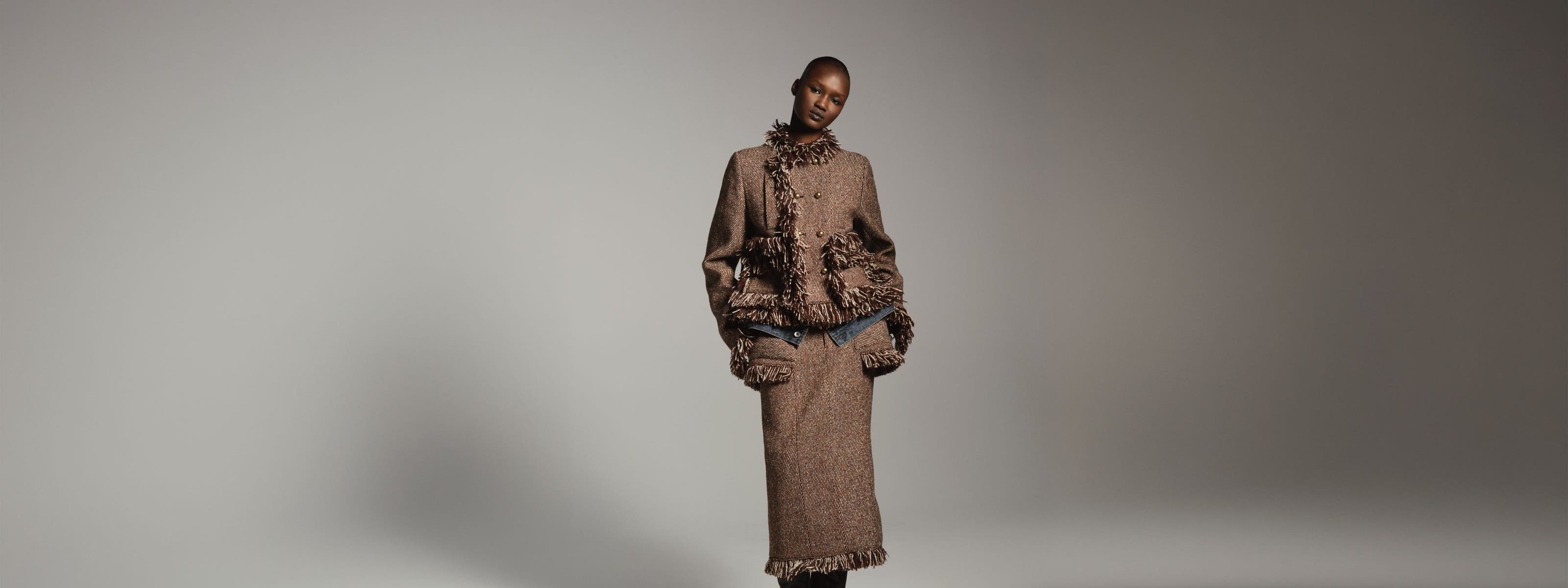 Woman wearing a textured brown fringed jacket with a matching long skirt and tall black boots, standing in a dramatically lit studio setting