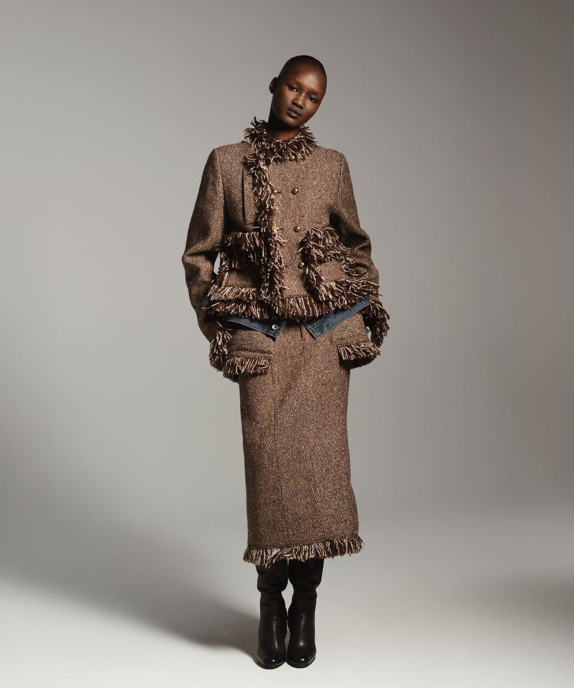 Woman wearing a textured brown fringed jacket with a matching long skirt and tall black boots, standing in a dramatically lit studio setting