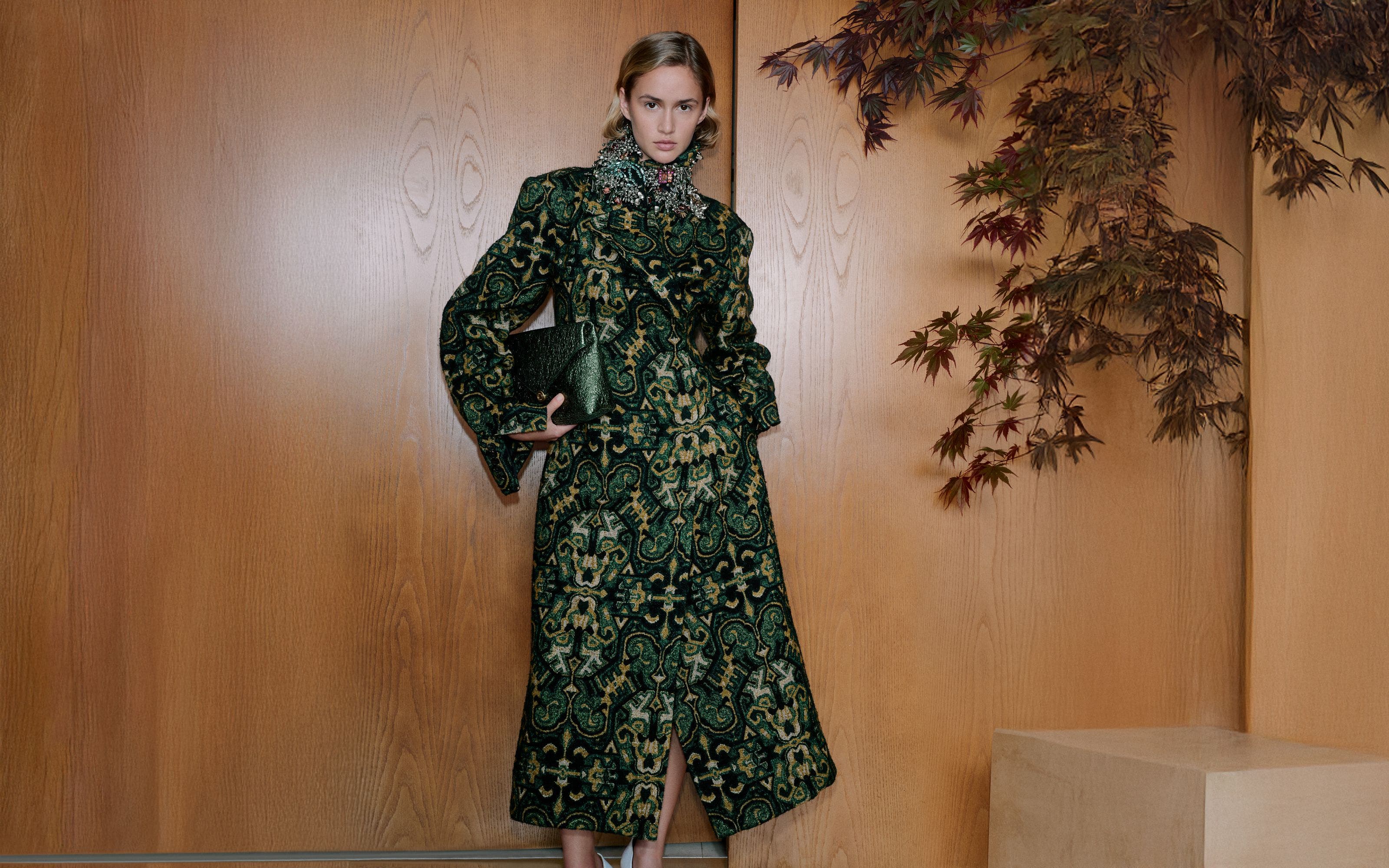 A woman wearing a patterned coat and sparkly collar necklace holding a handbag.