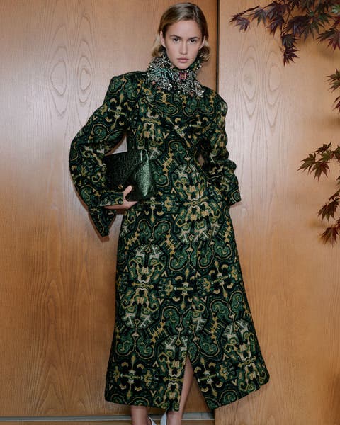 A woman wearing a patterned coat and sparkly collar necklace holding a handbag.