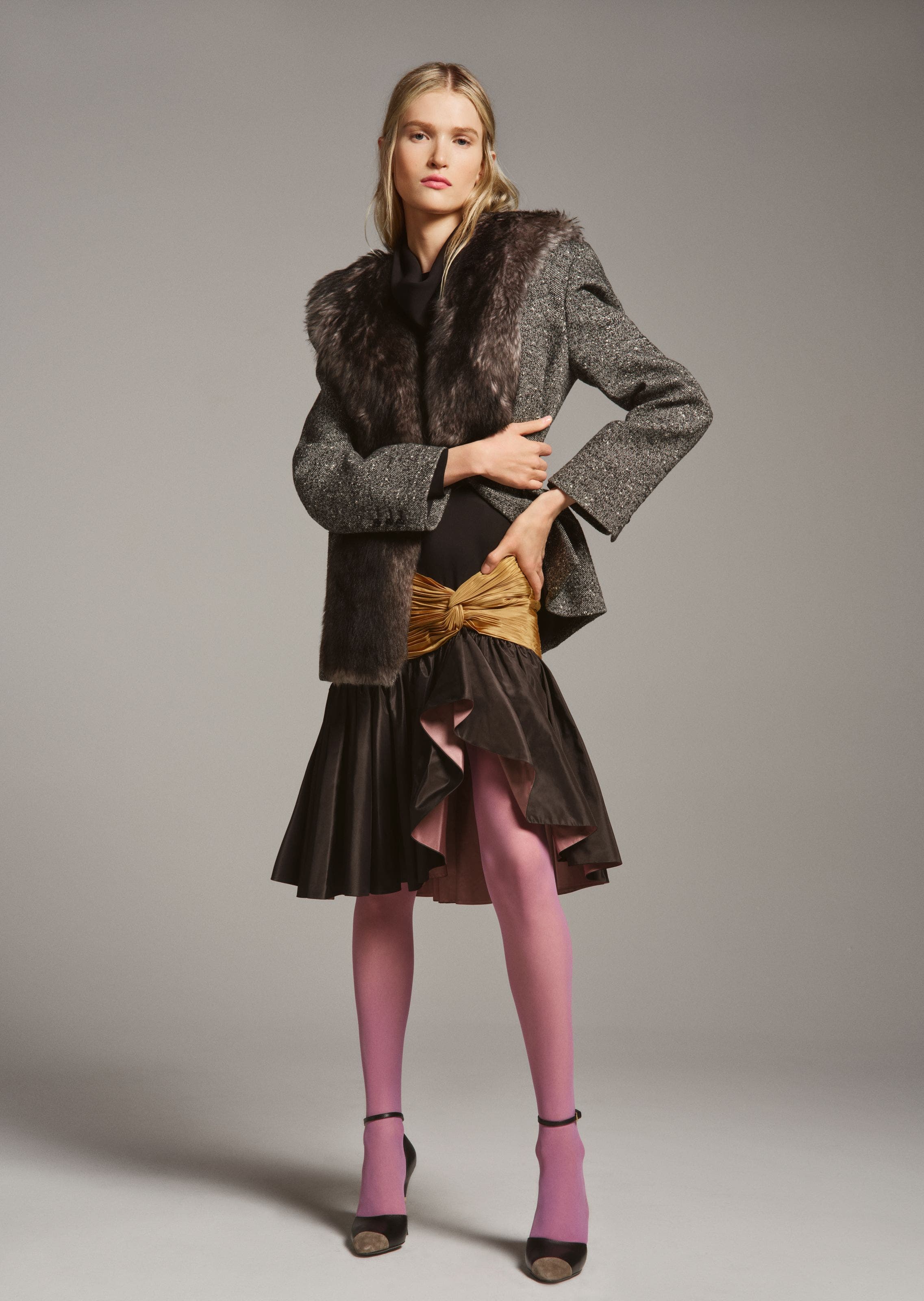 Woman wearing a gray tweed jacket with a fur collar and cuffs, paired with a mustard bow-front skirt over a black ruffled underlayer with pink lining