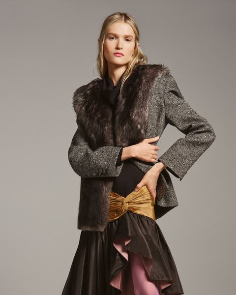 Woman wearing a gray tweed jacket with a fur collar and cuffs, paired with a mustard bow-front skirt over a black ruffled underlayer with pink lining