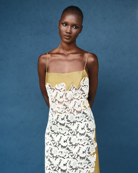Woman wearing a spaghetti-strap slip dress in ivory floral lace over a mustard-yellow underlay, shown against a blue background.