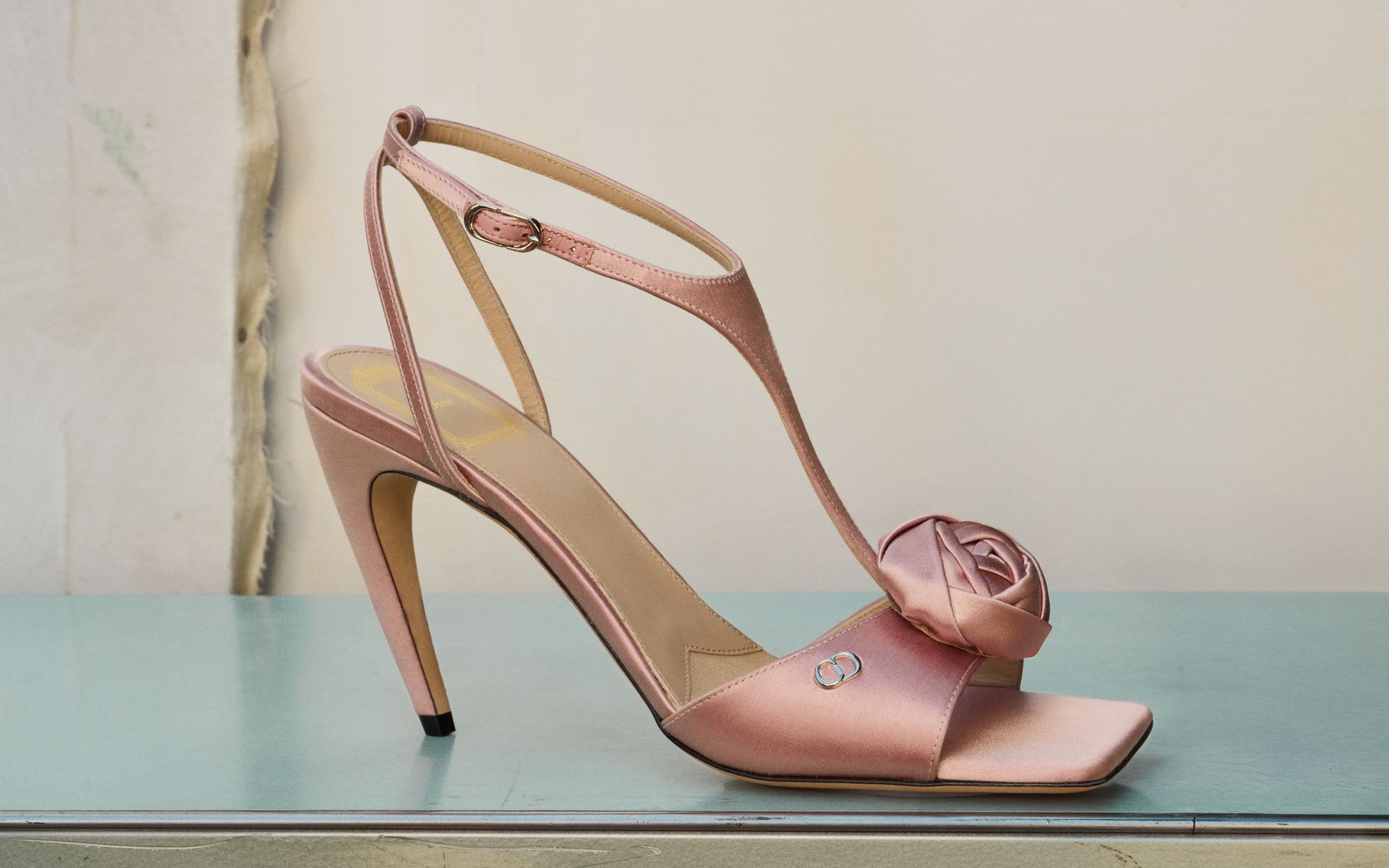Pink satin heeled sandal with an asymmetrical ankle strap and rosette detail, shown on a pastel pedestal