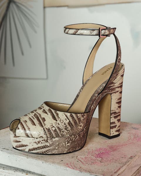 Snake-print peep-toe platform sandal in beige and brown tones with an ankle strap and chunky block heel, styled on a distressed pink surface.