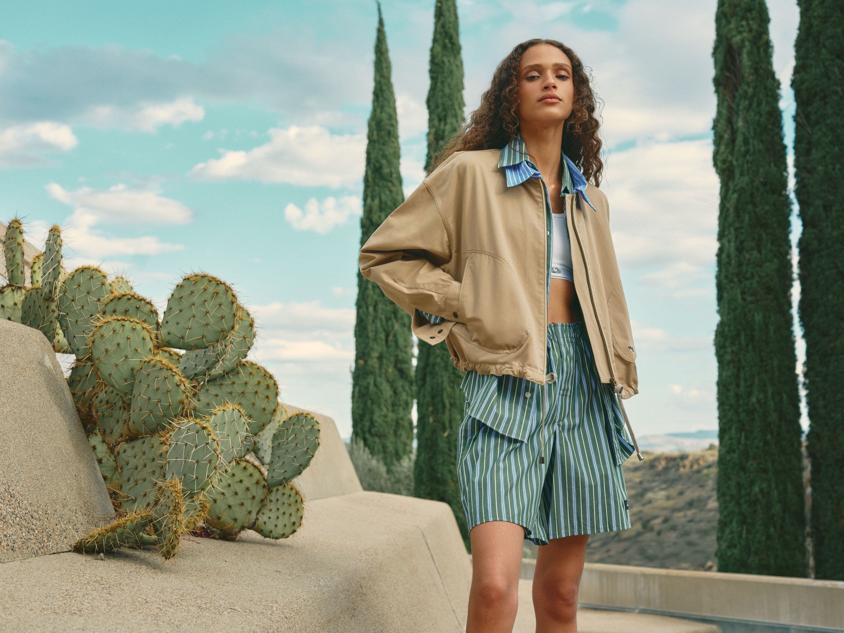 Woman wearing a parachute jacket with striped collar, sports bra, button-up shirt and boxer shorts.