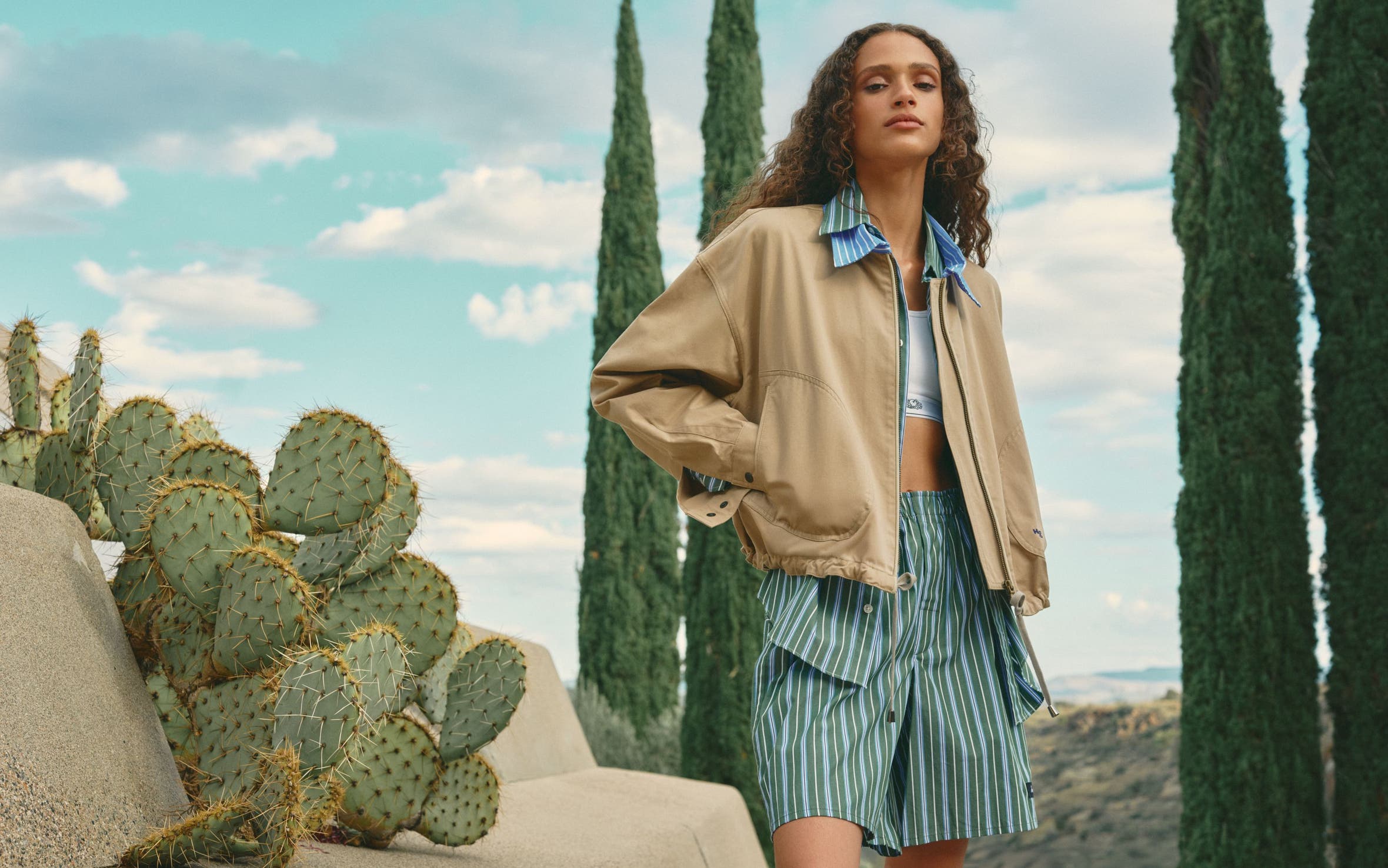 Woman wearing a parachute jacket with striped collar, sports bra, button-up shirt and boxer shorts.