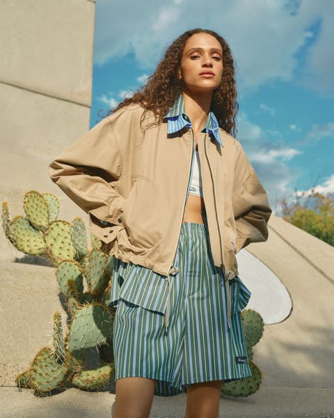 Woman wearing a khaki jacket, Oxford shirt, sports bra and boxer shorts.