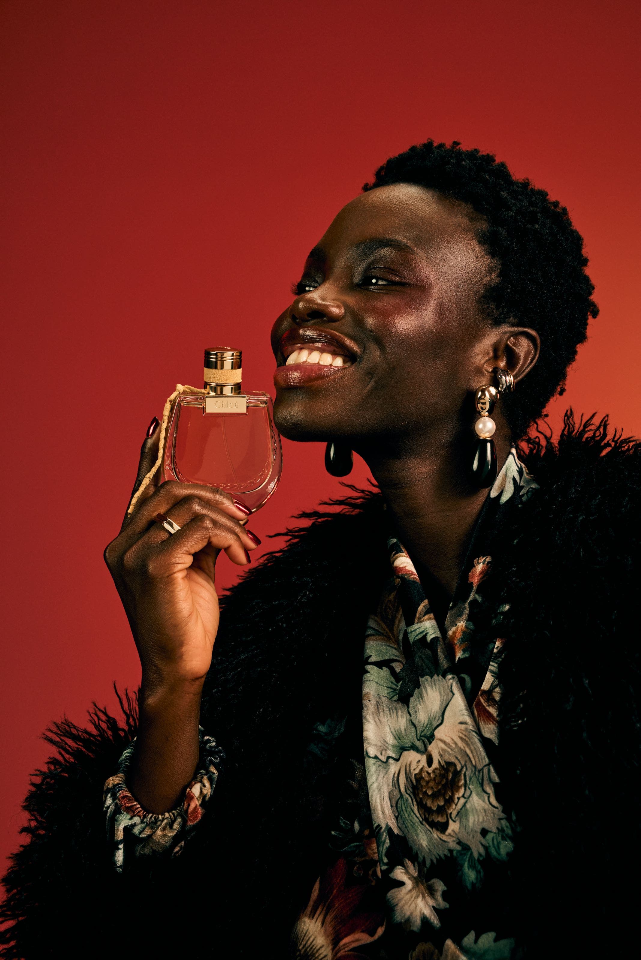 Model holding a Chloé perfume bottle while wearing a black feather coat over a floral top with mixed earrings.