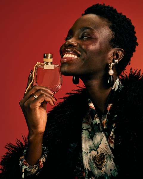 Model holding a Chloé perfume bottle while wearing a black feather coat over a floral top with mixed earrings.