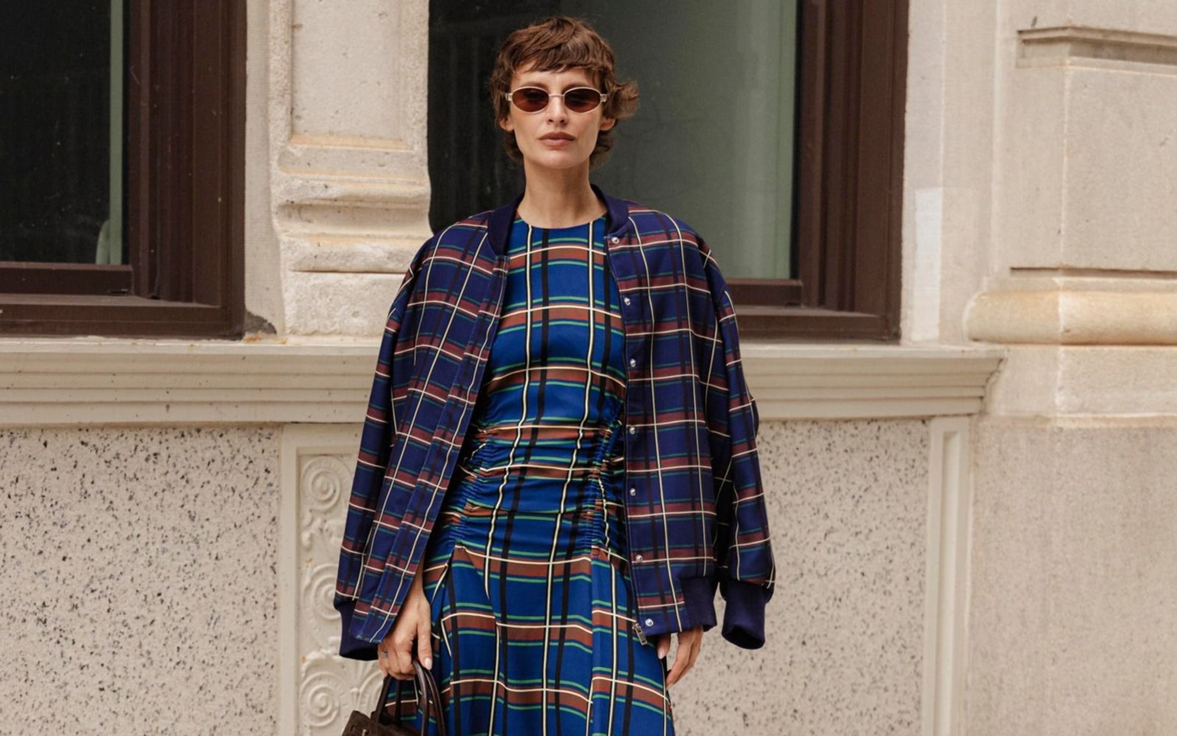 Woman wearing a navy plaid coat over a matching plaid midi dress, carrying a dark brown top-handle bag and wearing brown knee-high boots, standing on a city sidewalk
