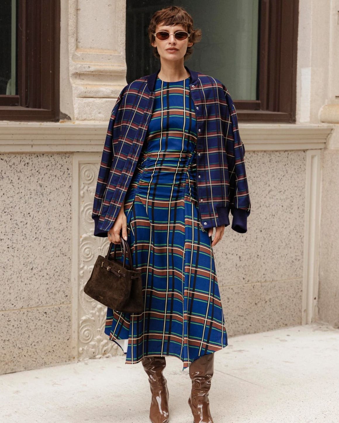 Woman wearing a navy plaid coat over a matching plaid midi dress, carrying a dark brown top-handle bag and wearing brown knee-high boots, standing on a city sidewalk