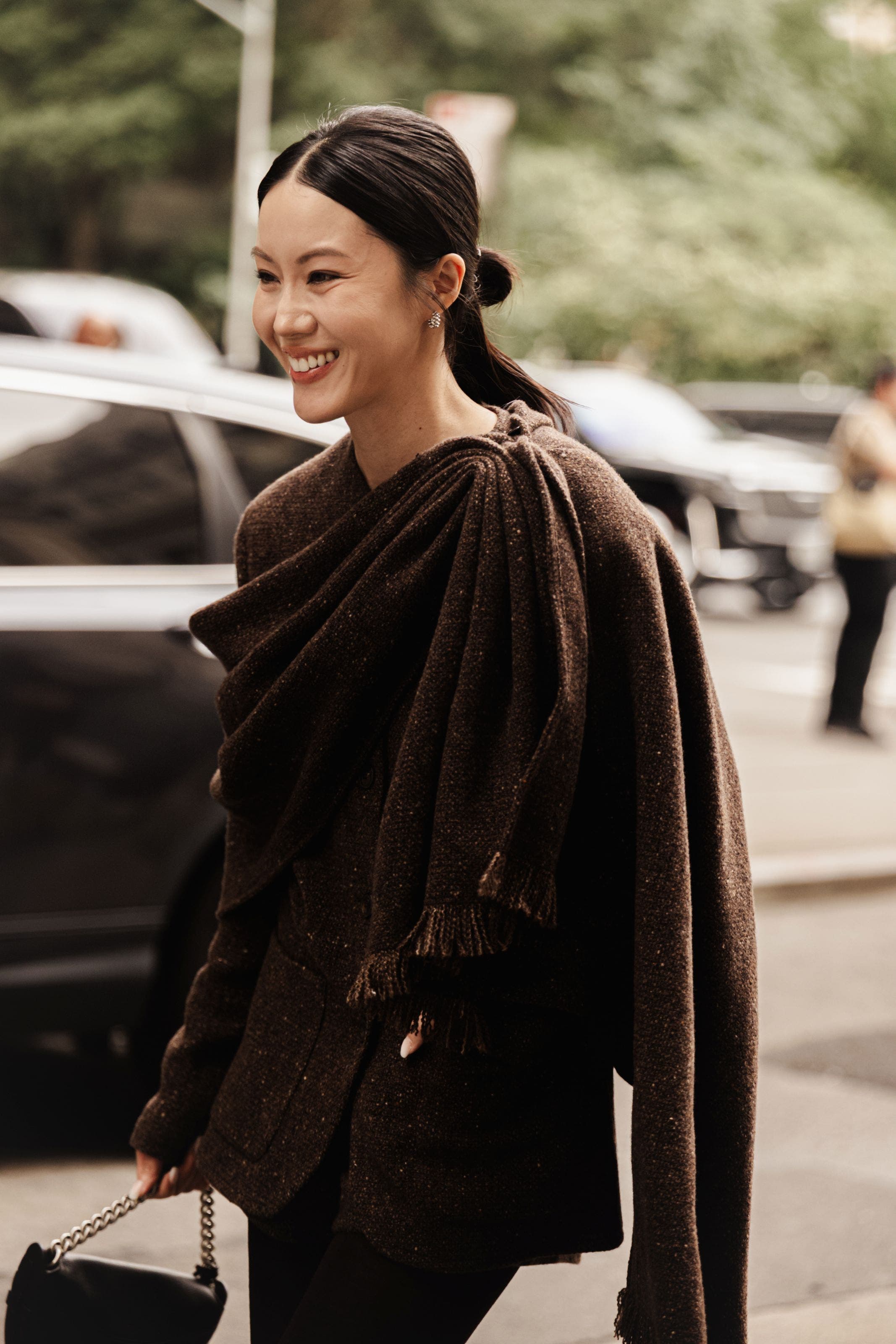 Woman smiling on a city street wearing a brown draped scarf and blazer, carrying a small black shoulder bag