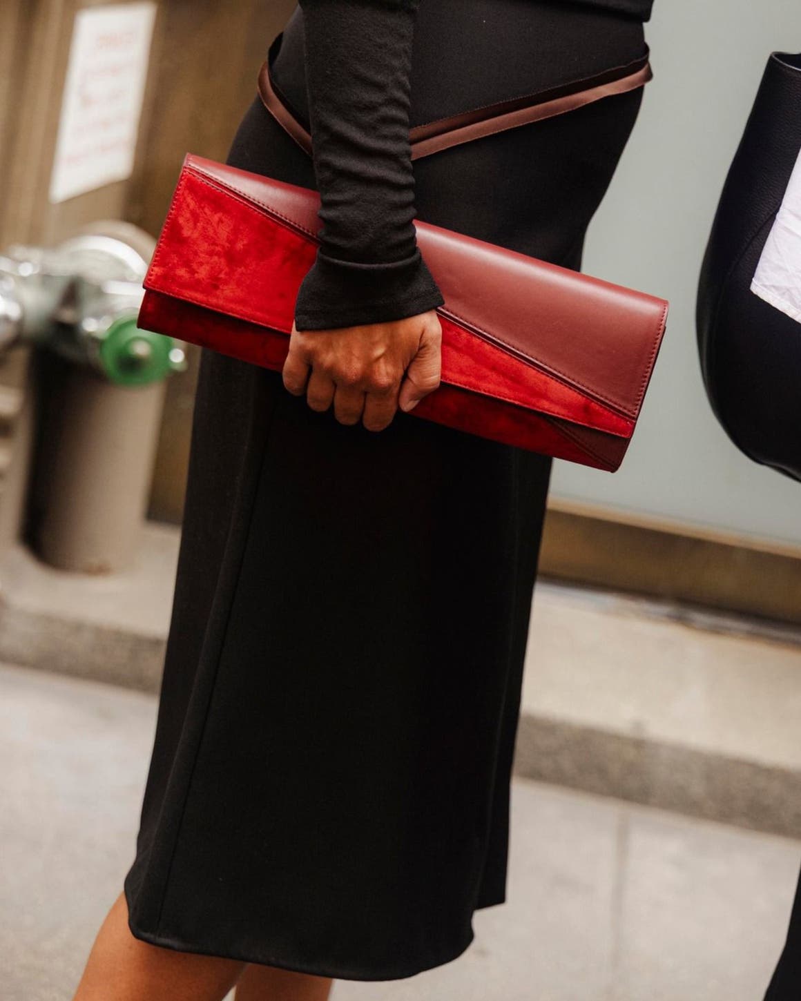 Close-up of a woman in a black dress holding a red and burgundy leather clutch, street style setting