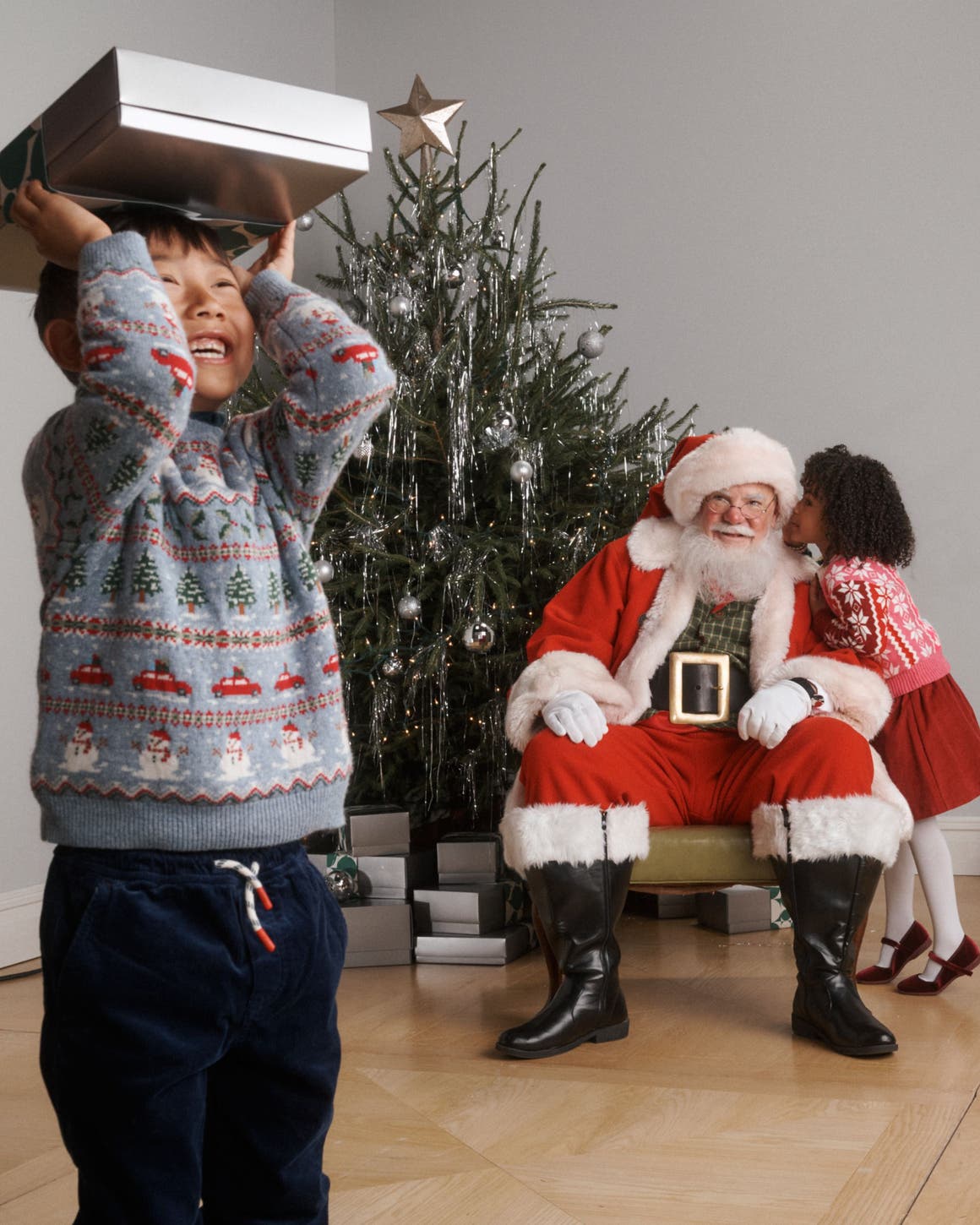 A kid holding up a gift box with Santa in the back.