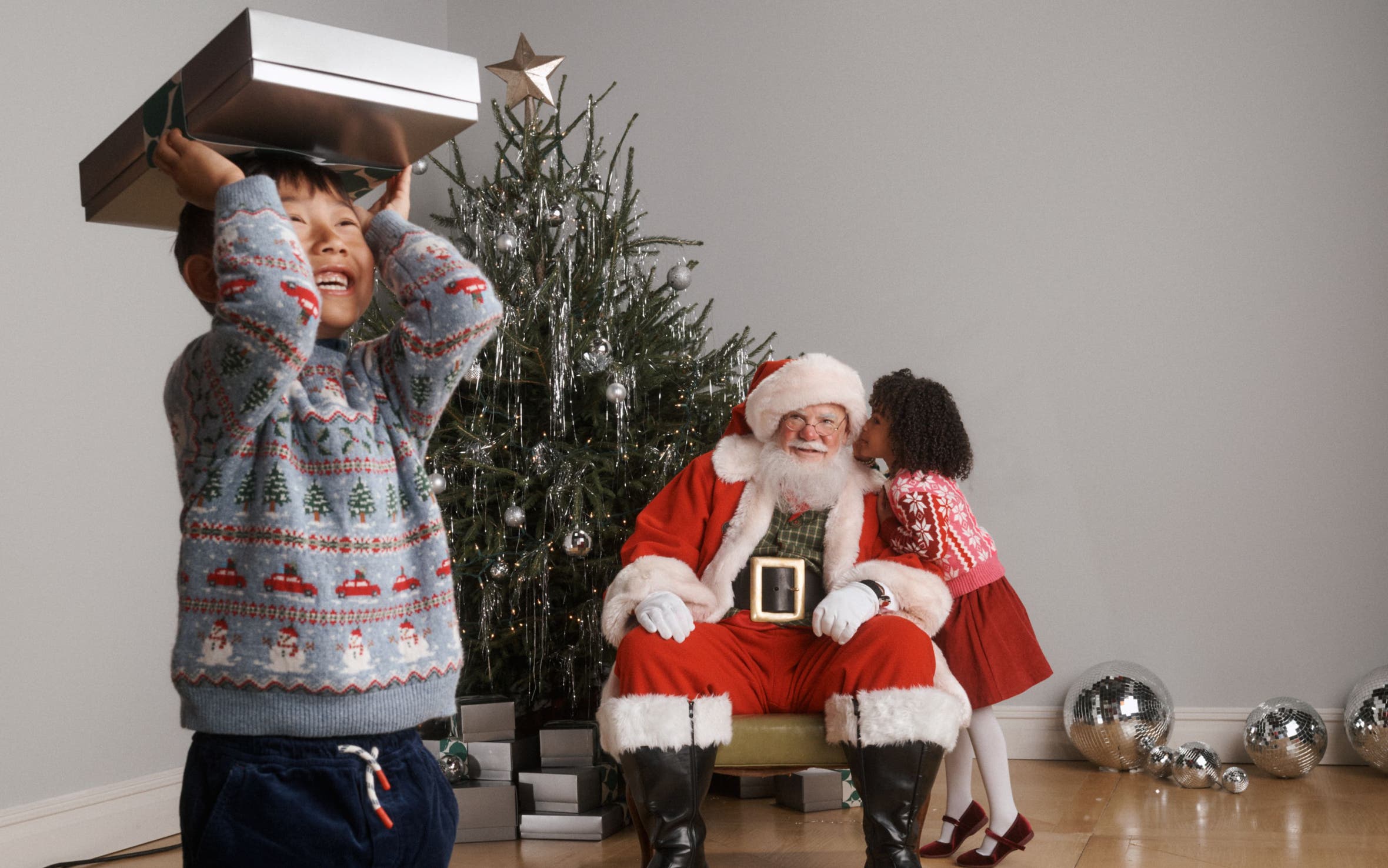 A kid holding up a gift box with Santa in the back.