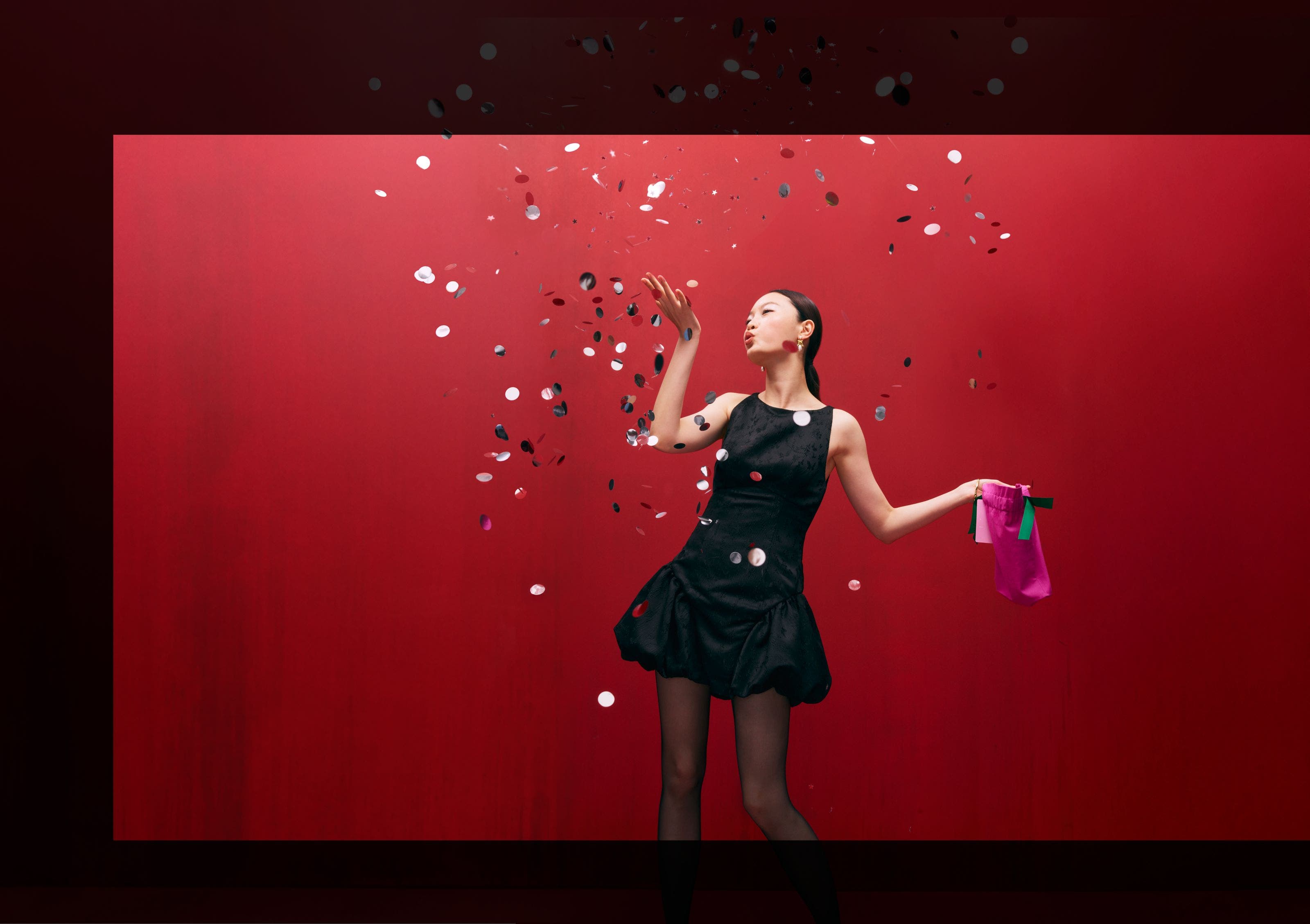 A woman in a black balloon minidress blowing pieces of confetti.