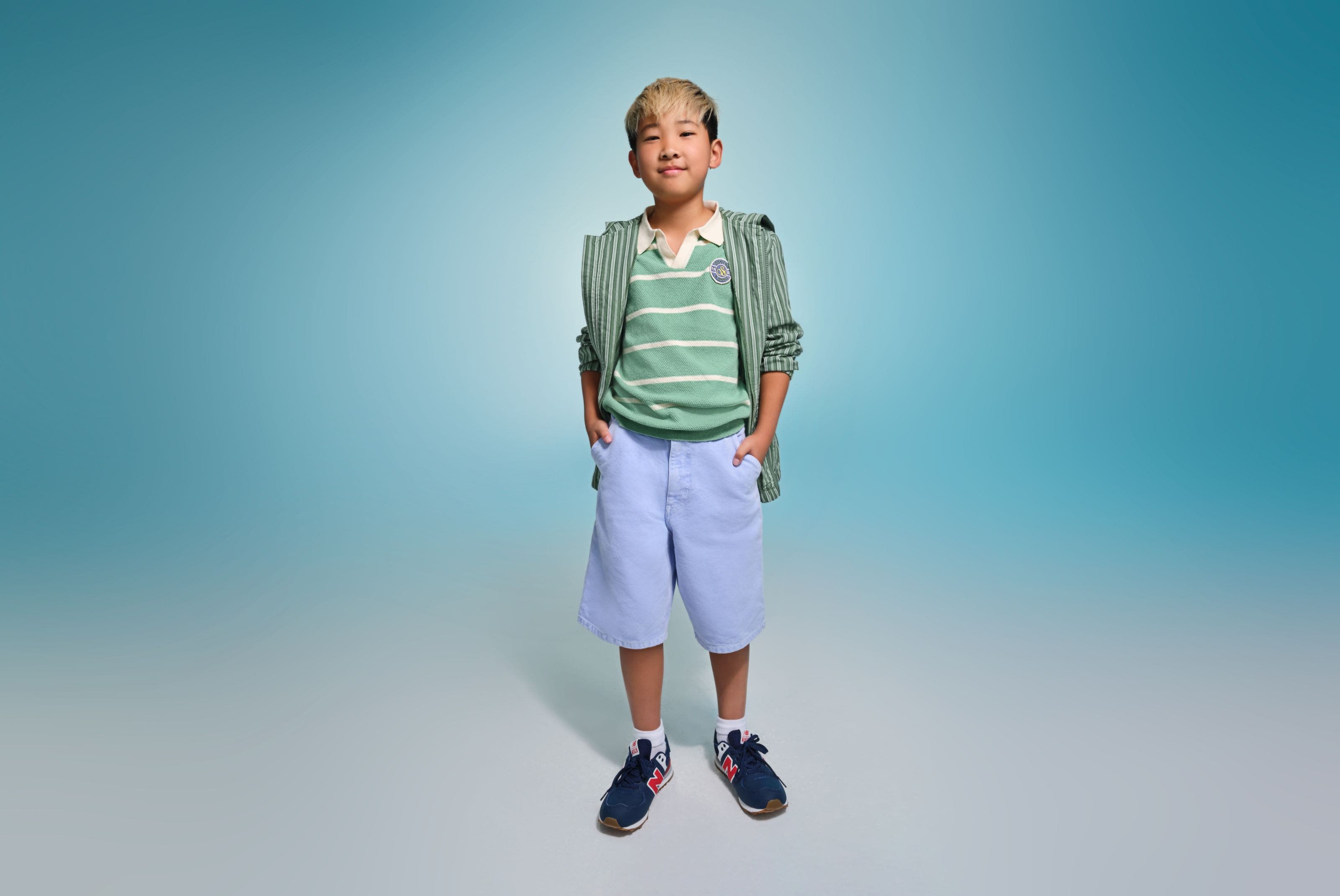A boy wearing a green striped polo and jacket with purple shorts and navy sneakers.