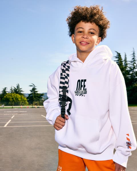 Boy in a white Off-White logo hoodie standing outdoors with trees and a parking lot in the background.