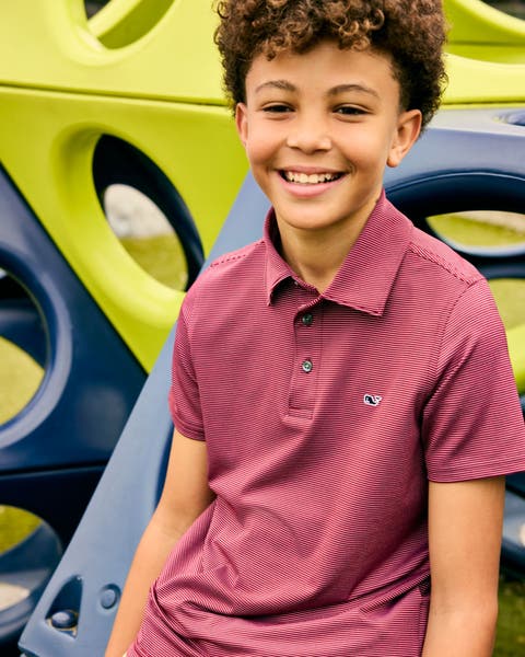 Boy in a red Vineyard Vines polo shirt smiling in front of a yellow and navy playground structure.