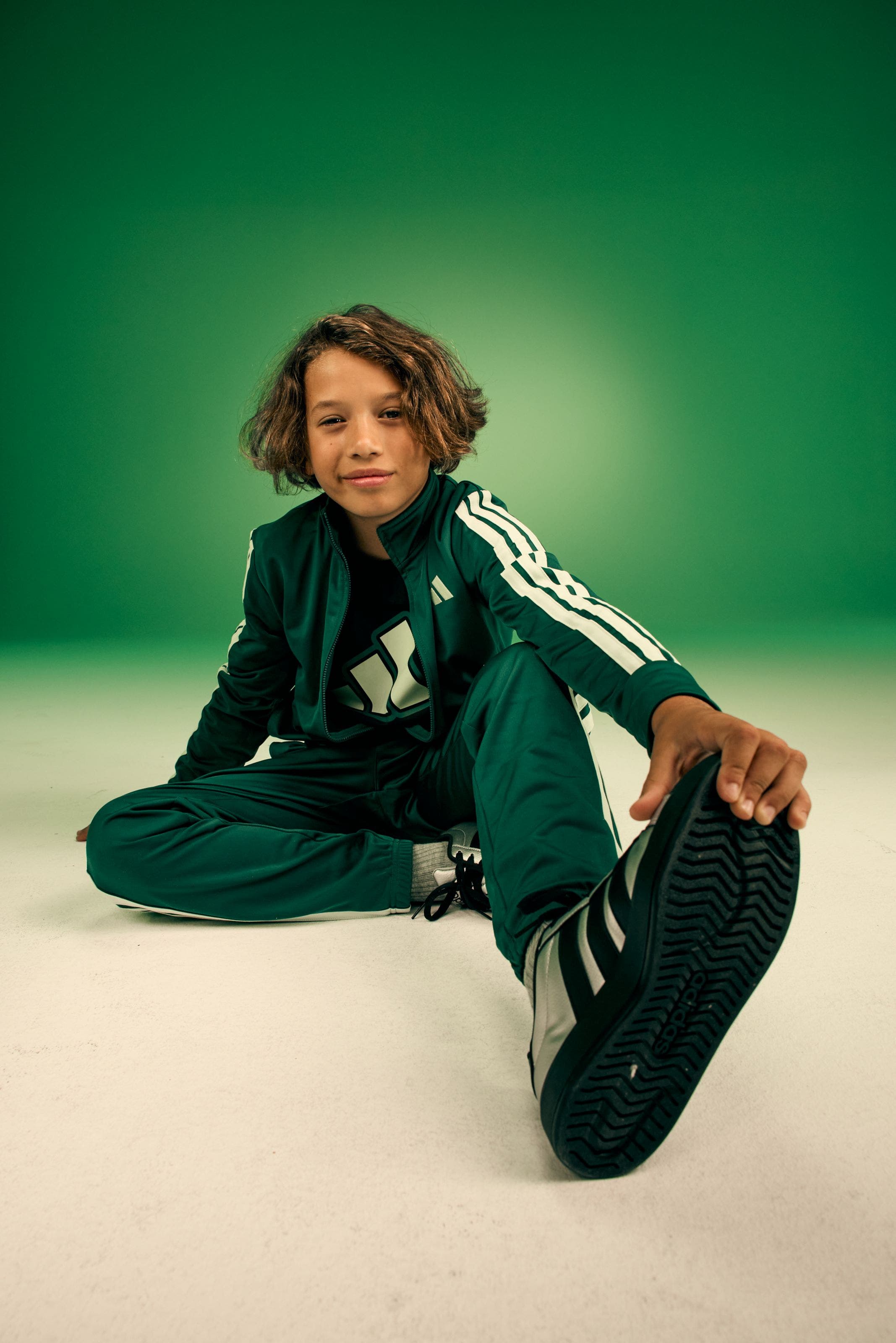 Child model wearing a dark green Adidas track suit with white three-stripe detail and matching green sneakers.