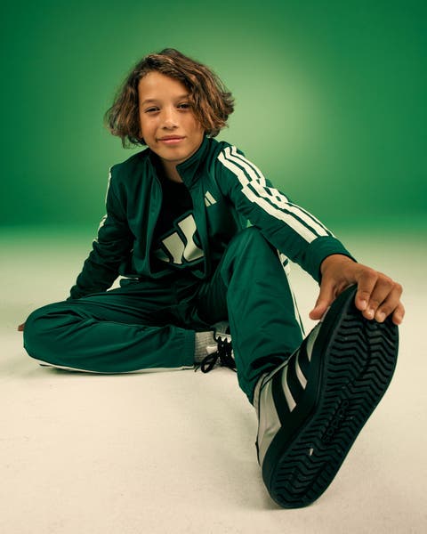 Child model wearing a dark green Adidas track suit with white three-stripe detail and matching green sneakers.