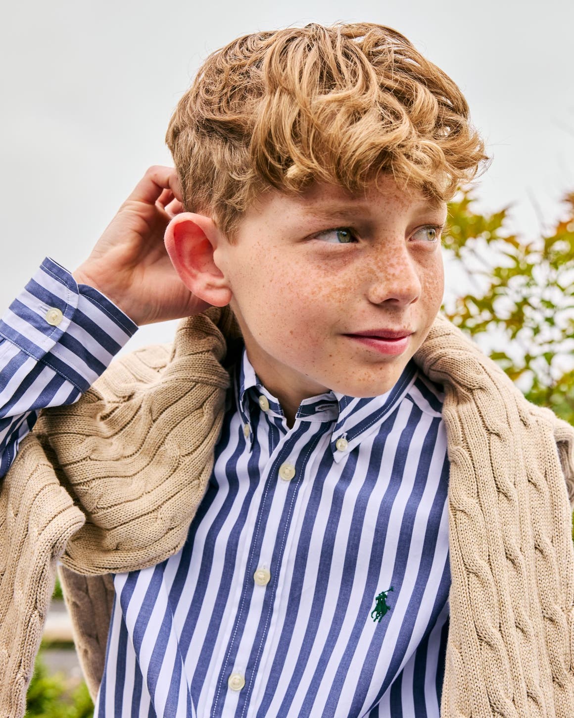 Boy wearing a Polo Ralph Lauren blue-and-white striped button-down shirt layered under a cream cable-knit sweater.