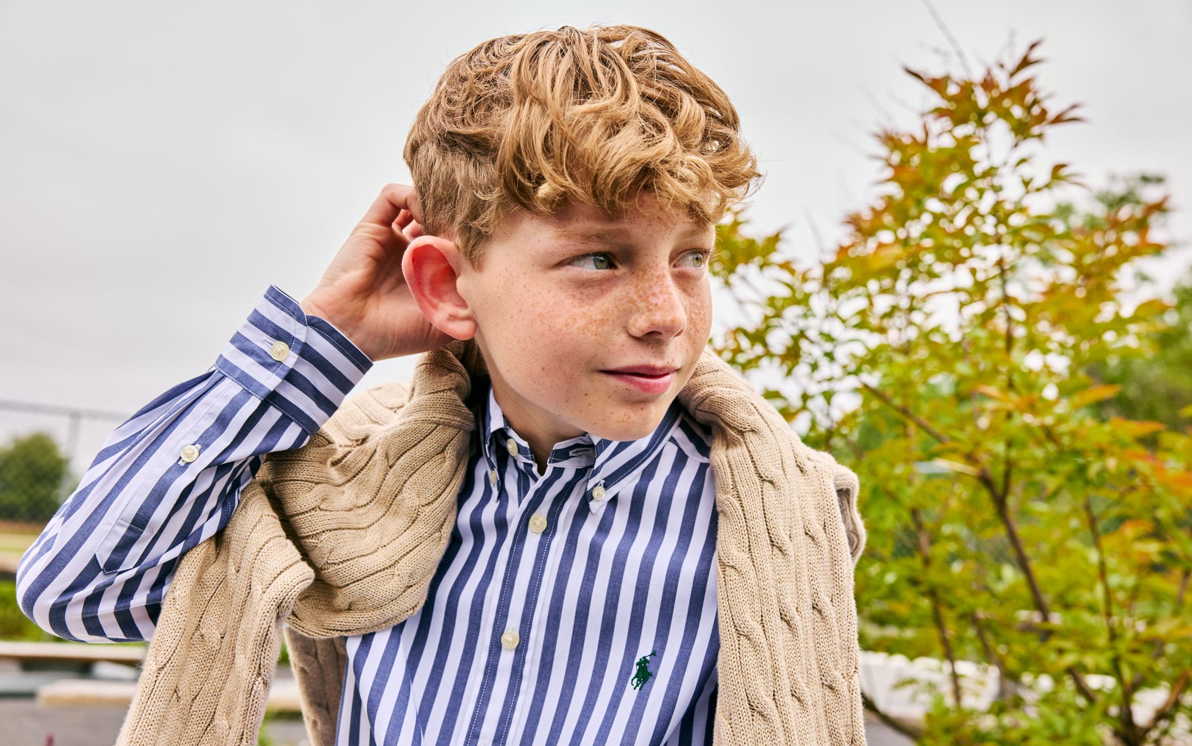 Boy wearing a Polo Ralph Lauren blue-and-white striped button-down shirt layered under a cream cable-knit sweater.