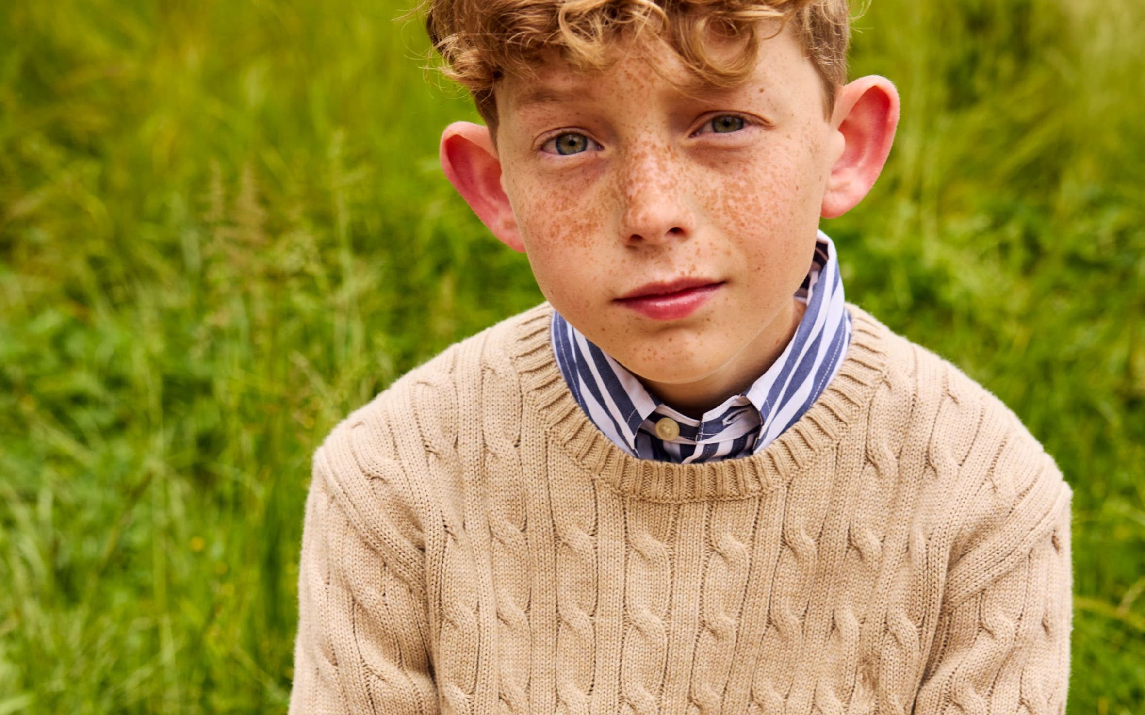 Boy in a tan cable-knit sweater over a blue and white striped collared shirt.