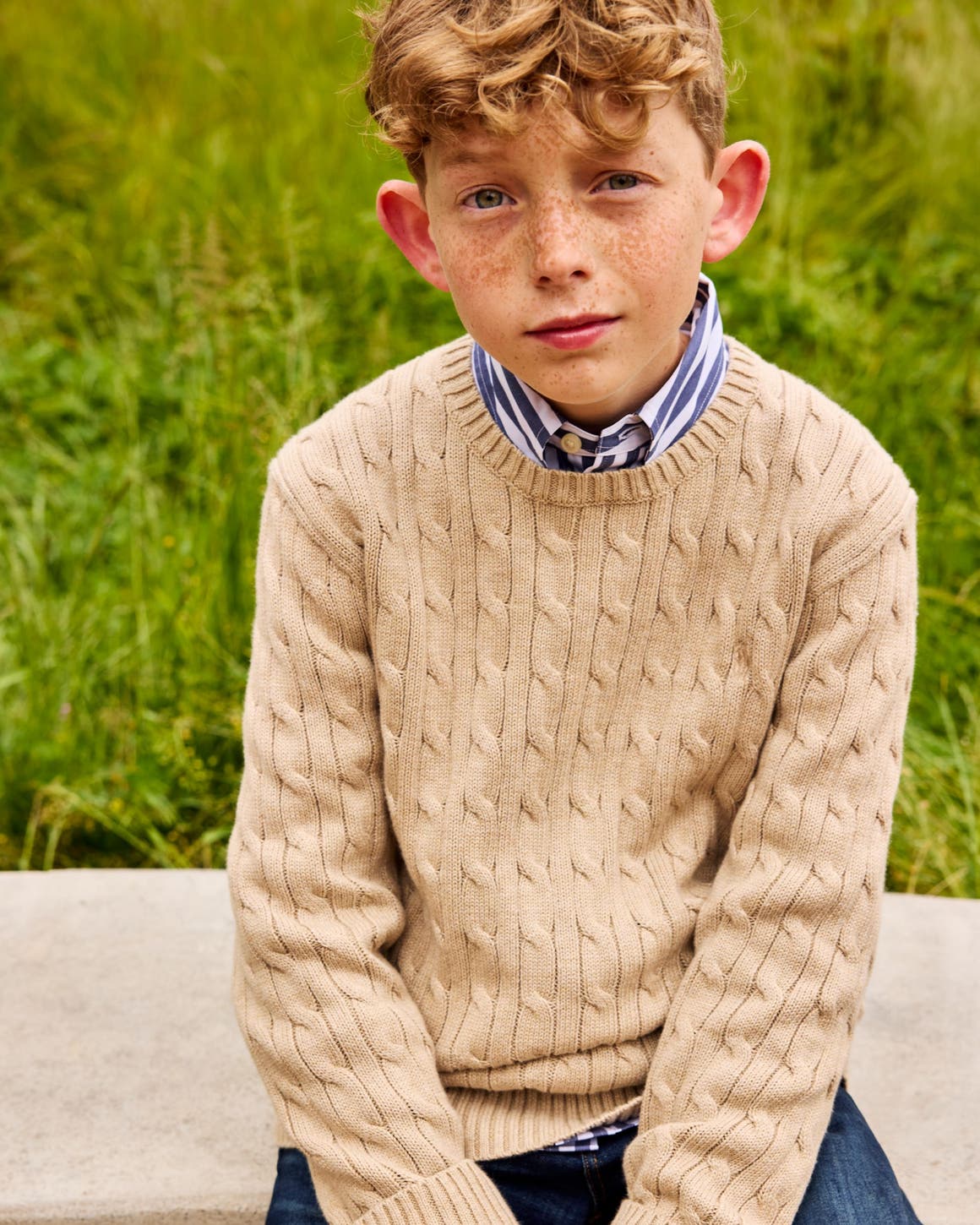 Boy in a tan cable-knit sweater over a blue and white striped collared shirt.