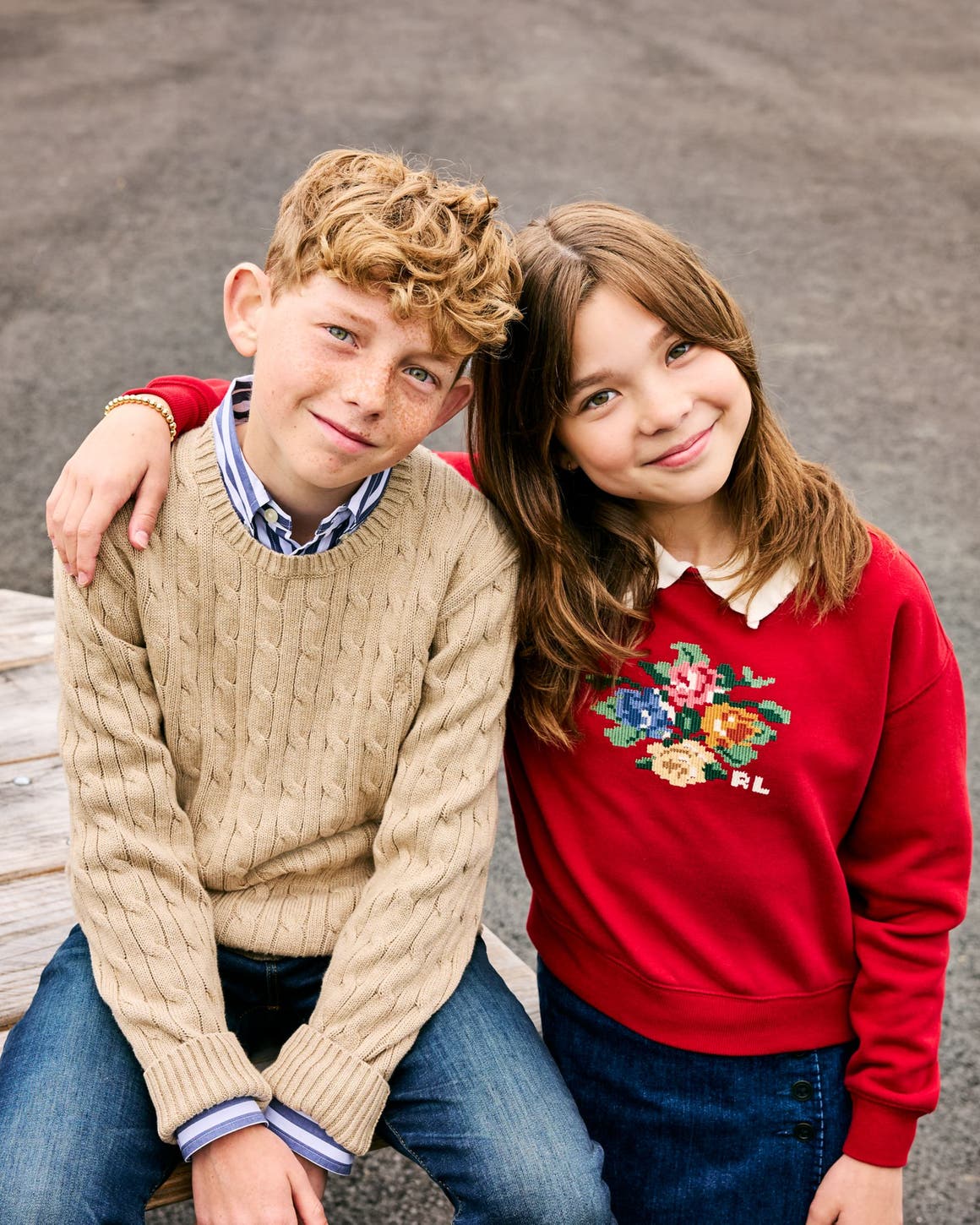 Two kids, a boy and a girl, sit together on a picnic table wearing cozy knit and cross-stitch sweaters.