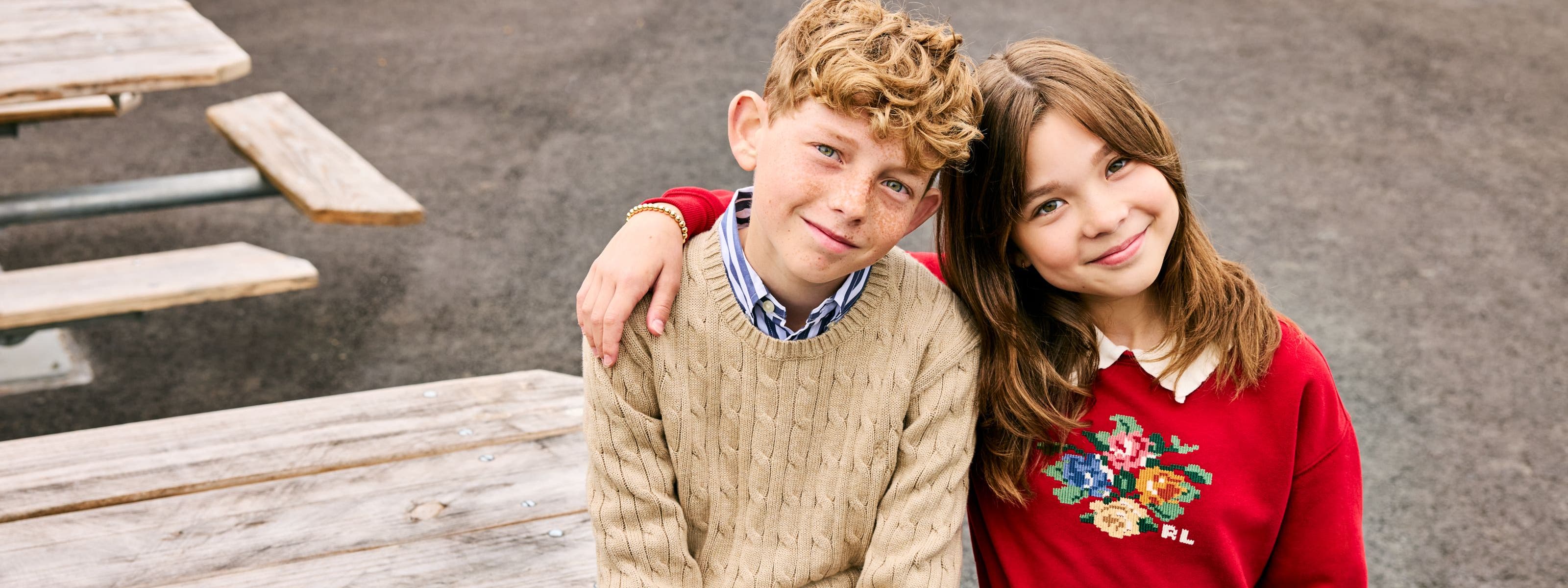 Two kids, a boy and a girl, sit together on a picnic table wearing cozy knit and cross-stitch sweaters.